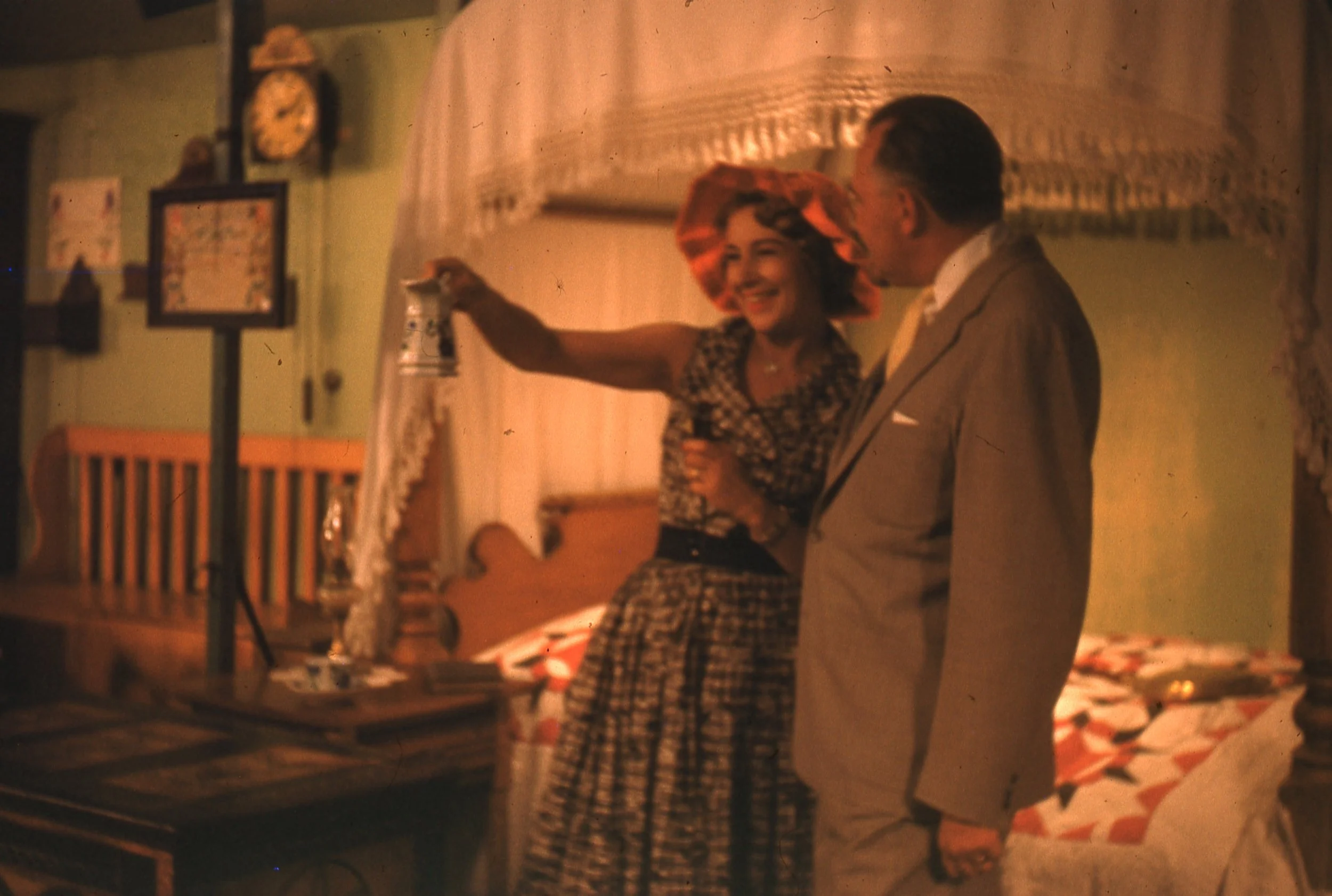 Man, wearing gray suit, and woman, wearing orange hat and holding microphone, standing on bedroom set with antique furniture.