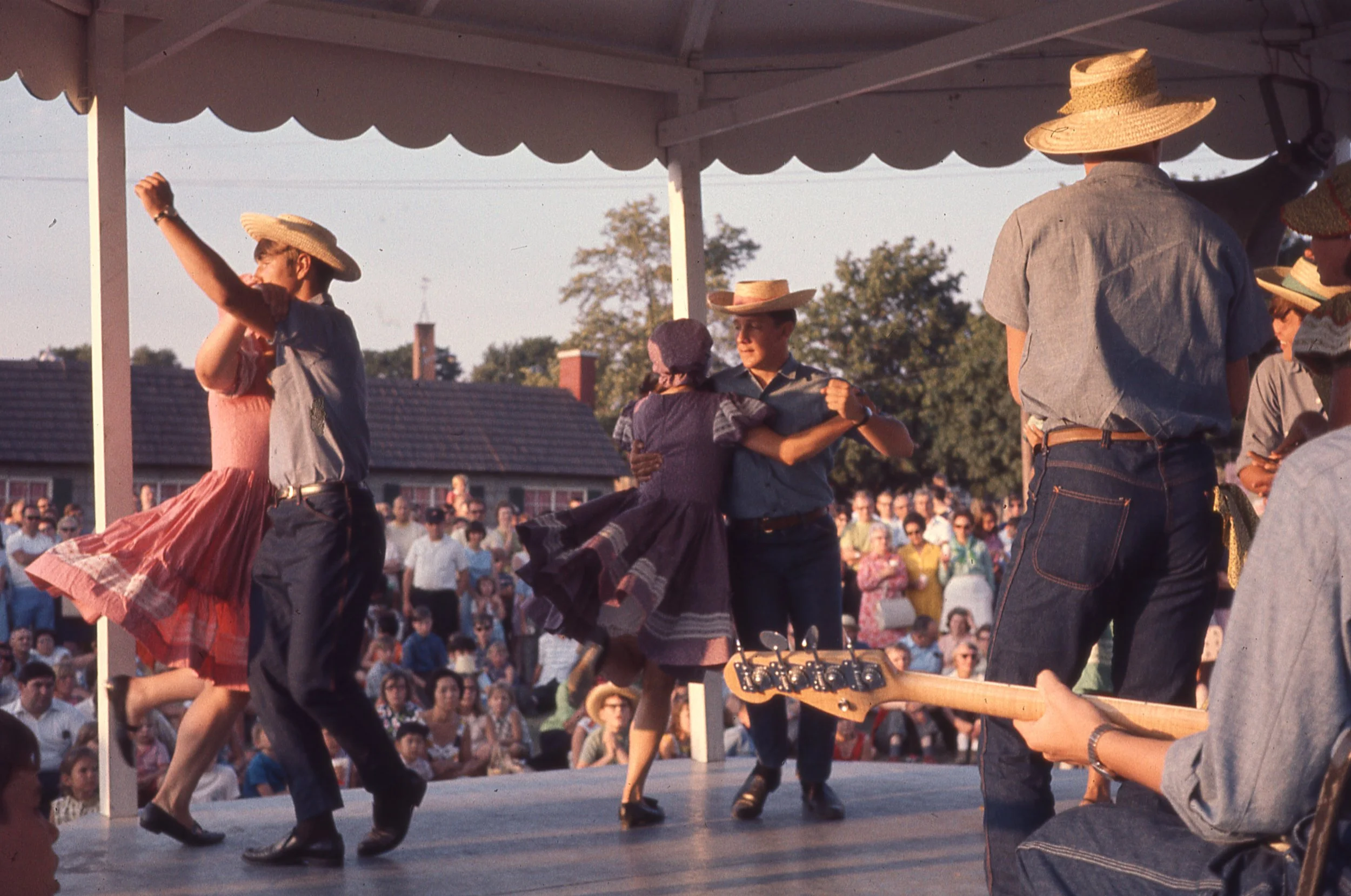 Pairs of women in matching dresses and bonnets and men in straw hats, dancing on covered outdoor stage.