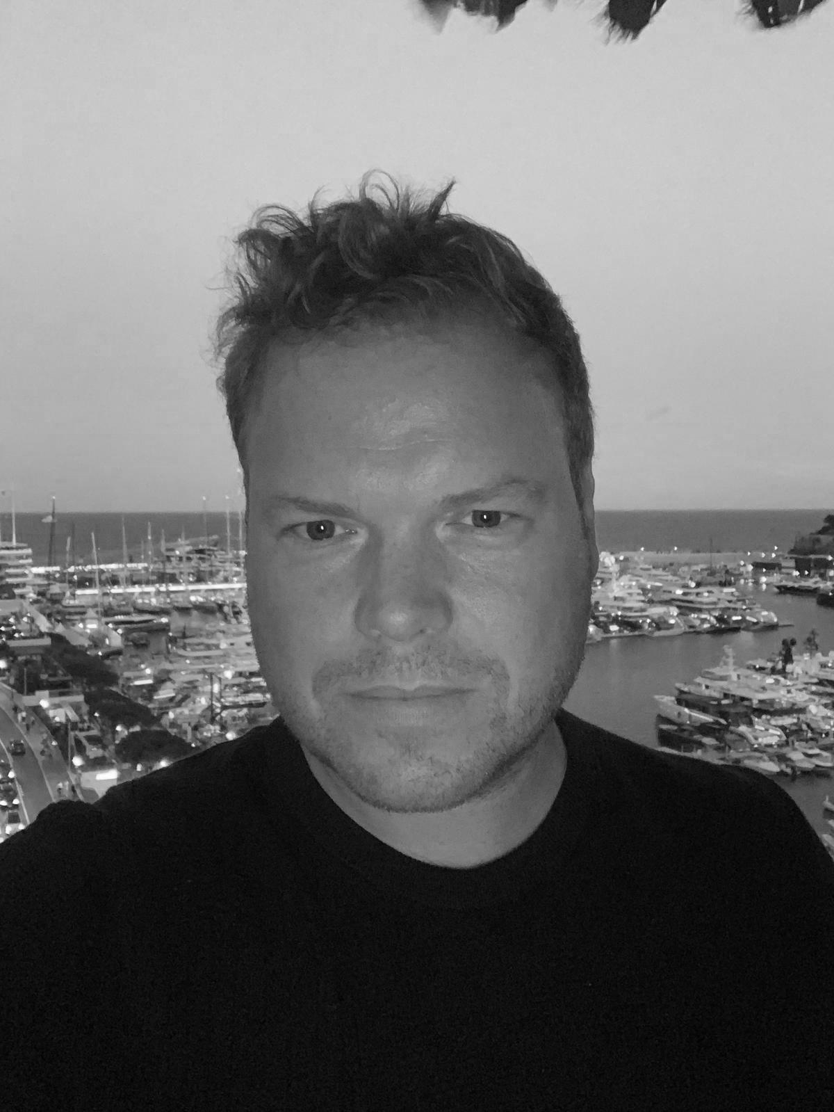 A man with short, curly hair and a slight beard taking a selfie in front of a marina filled with boats, with the ocean visible in the background, in black and white.