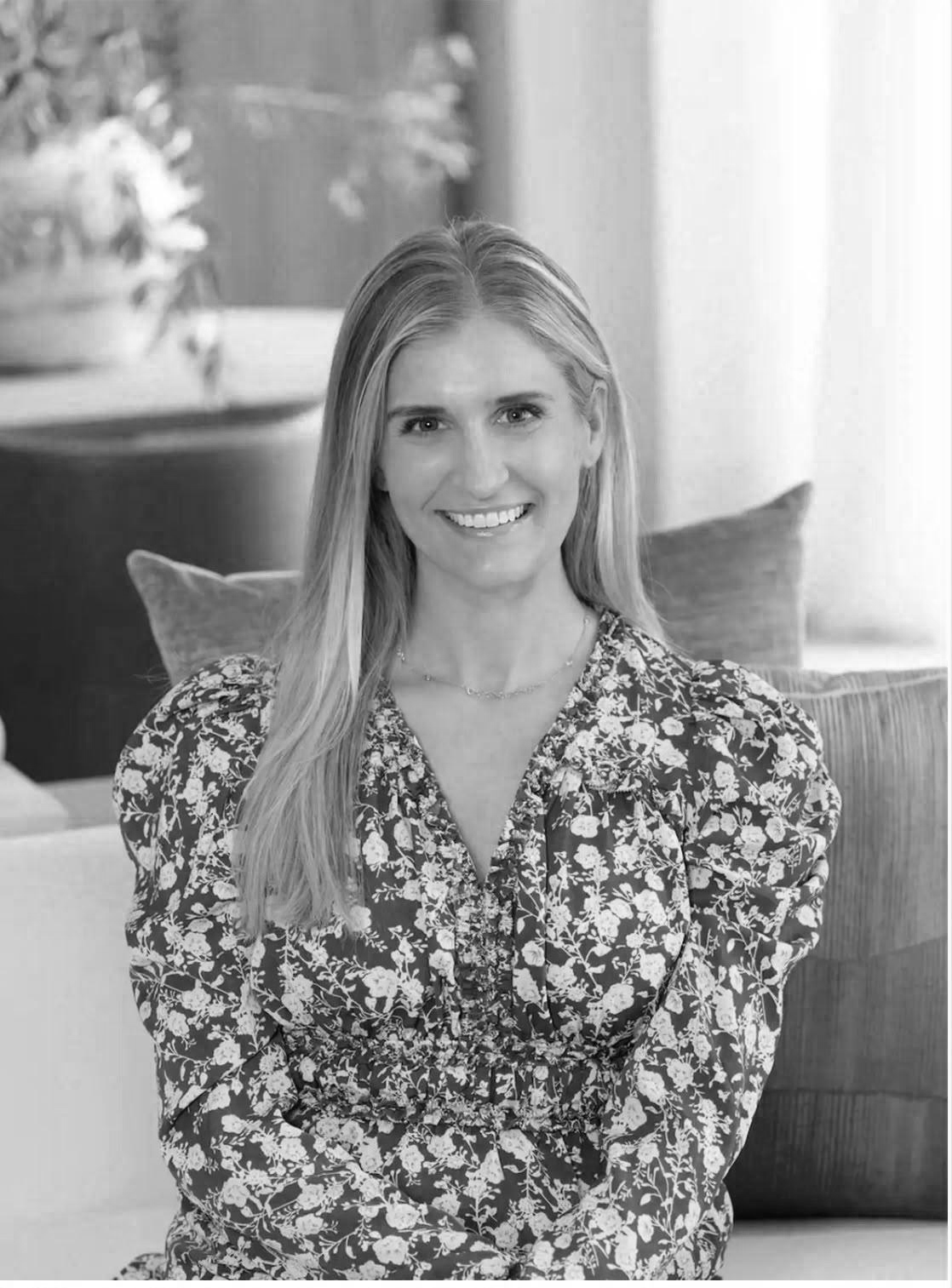 A smiling woman with long hair sitting on a couch in a living room with a potted plant in the background.