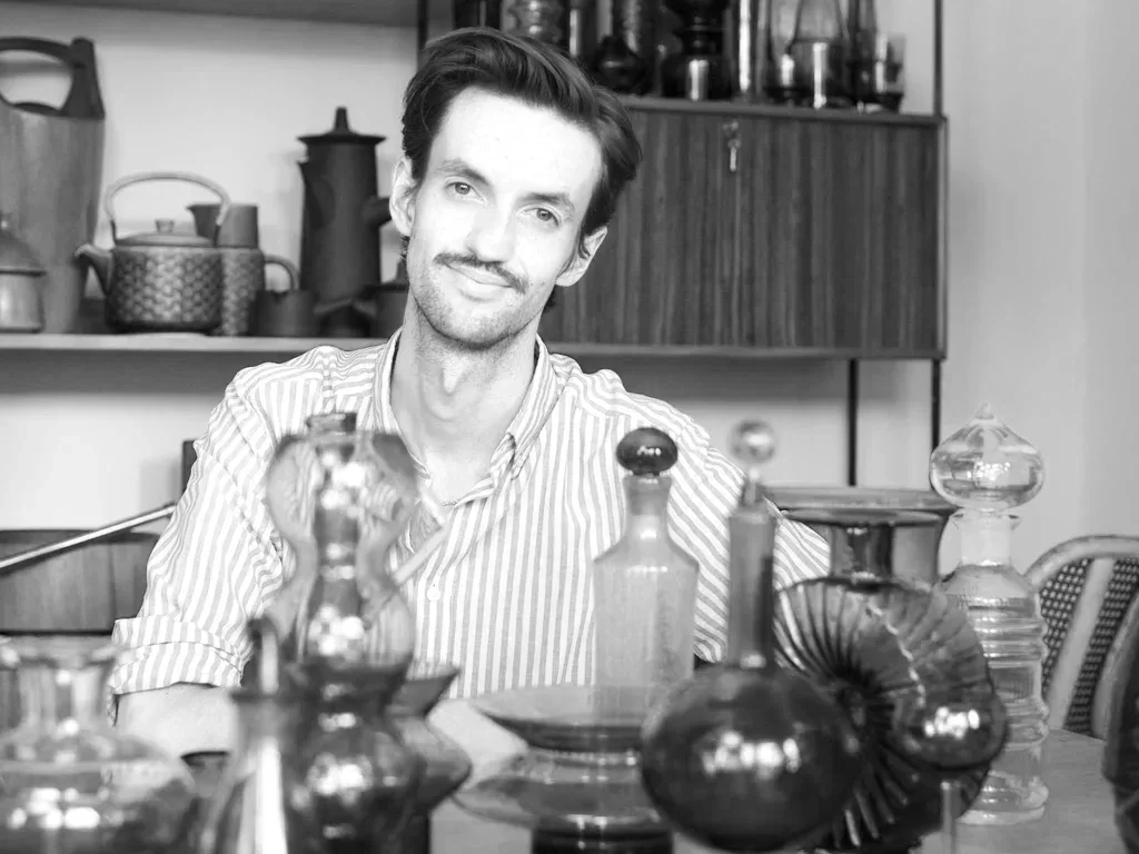 A man with dark hair, a mustache, and a slight smile sitting at a table covered with decorative glass bottles and vessels, with a kitchen shelf with teapots and jars in the background.