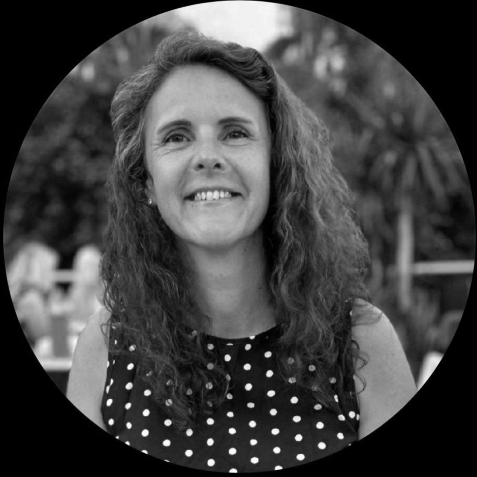 Black and white photo of a smiling woman with long, curly hair, wearing a polka dot top, outdoors with trees and a fence in the background.