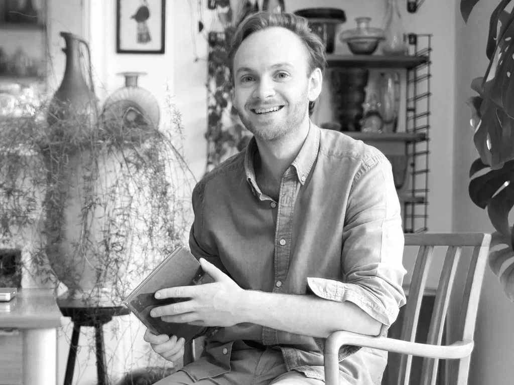 A man with light-colored hair and a beard smiling, sitting in a wooden chair in a room with plants and shelves.