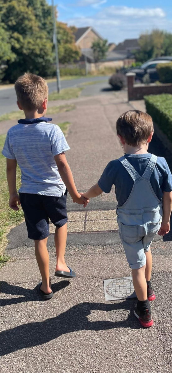 Two young boys walking on a sidewalk hand-in-hand in a suburban neighborhood, with houses and trees in the background, under a partly cloudy sky.