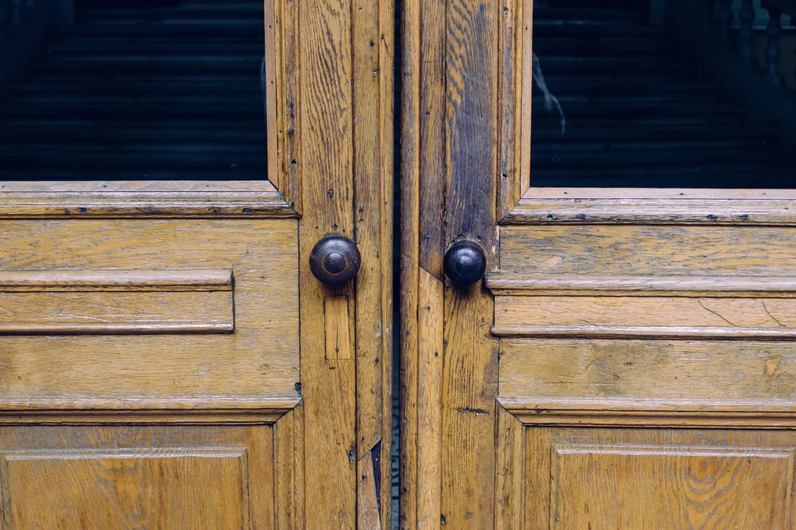 Close-up of wooden double doors with black round doorknobs, glass panels, and visible wear and scratches.