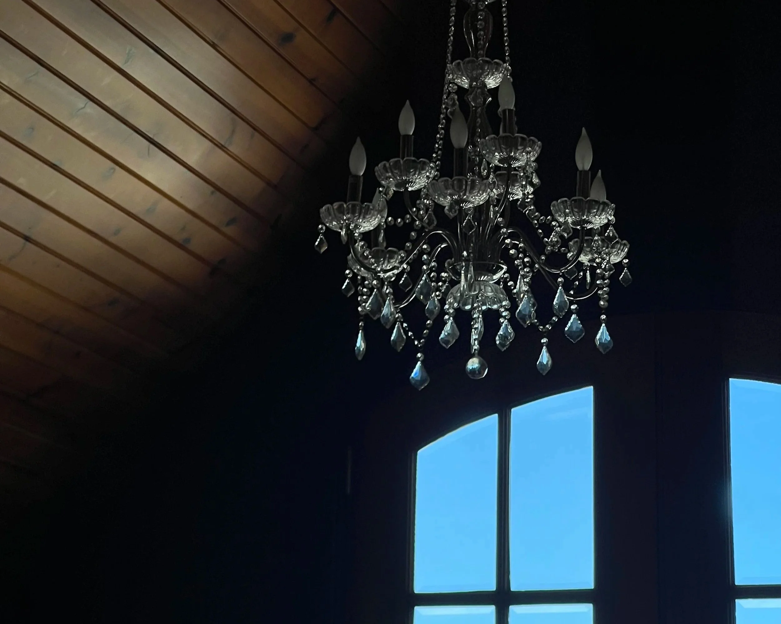 A chandelier with hanging crystals and candle-shaped lights. The ceiling features wooden beams and an area of sky blue window panes.