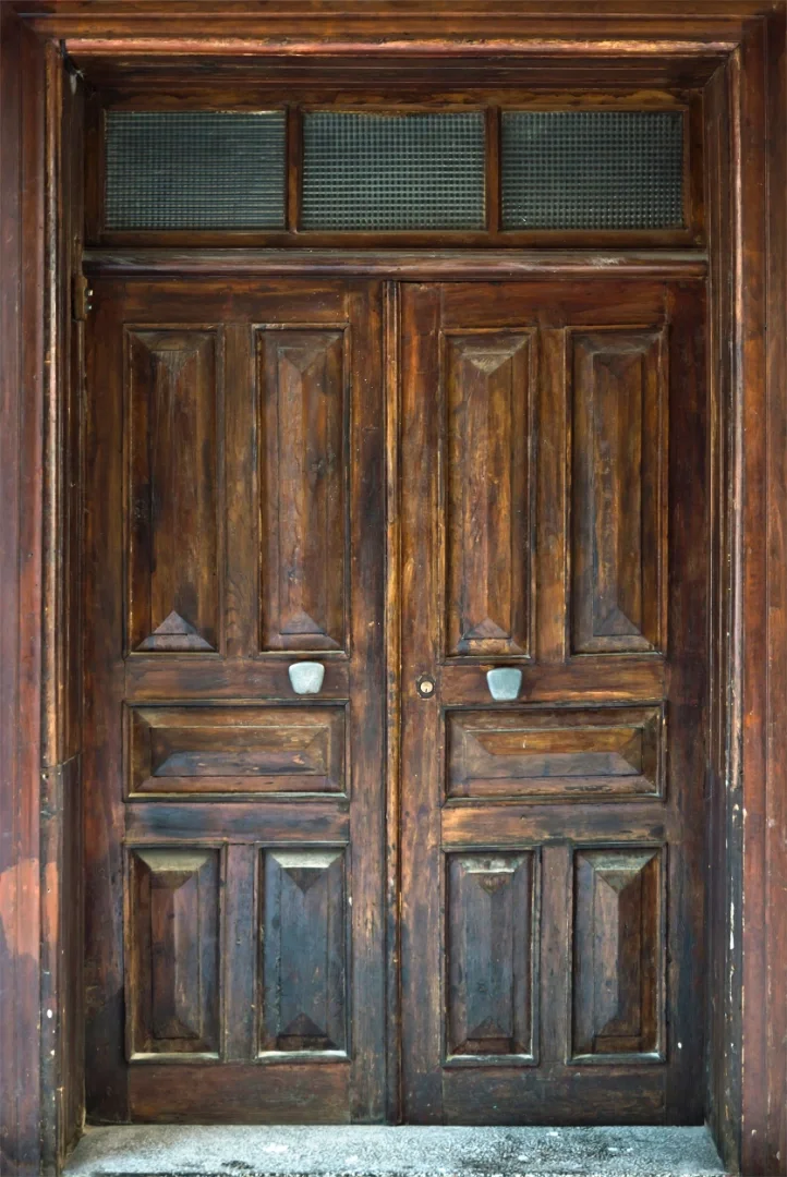 A pair of wooden doors with six rectangular panels on each door, set in a wooden frame. The doors have a small, conical door knocker on each, and a glass transom window above with four small glass panes.