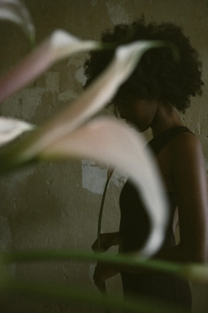 A woman with curly hair is seen in profile, standing against a textured wall, partially obscured by a plant in the foreground.