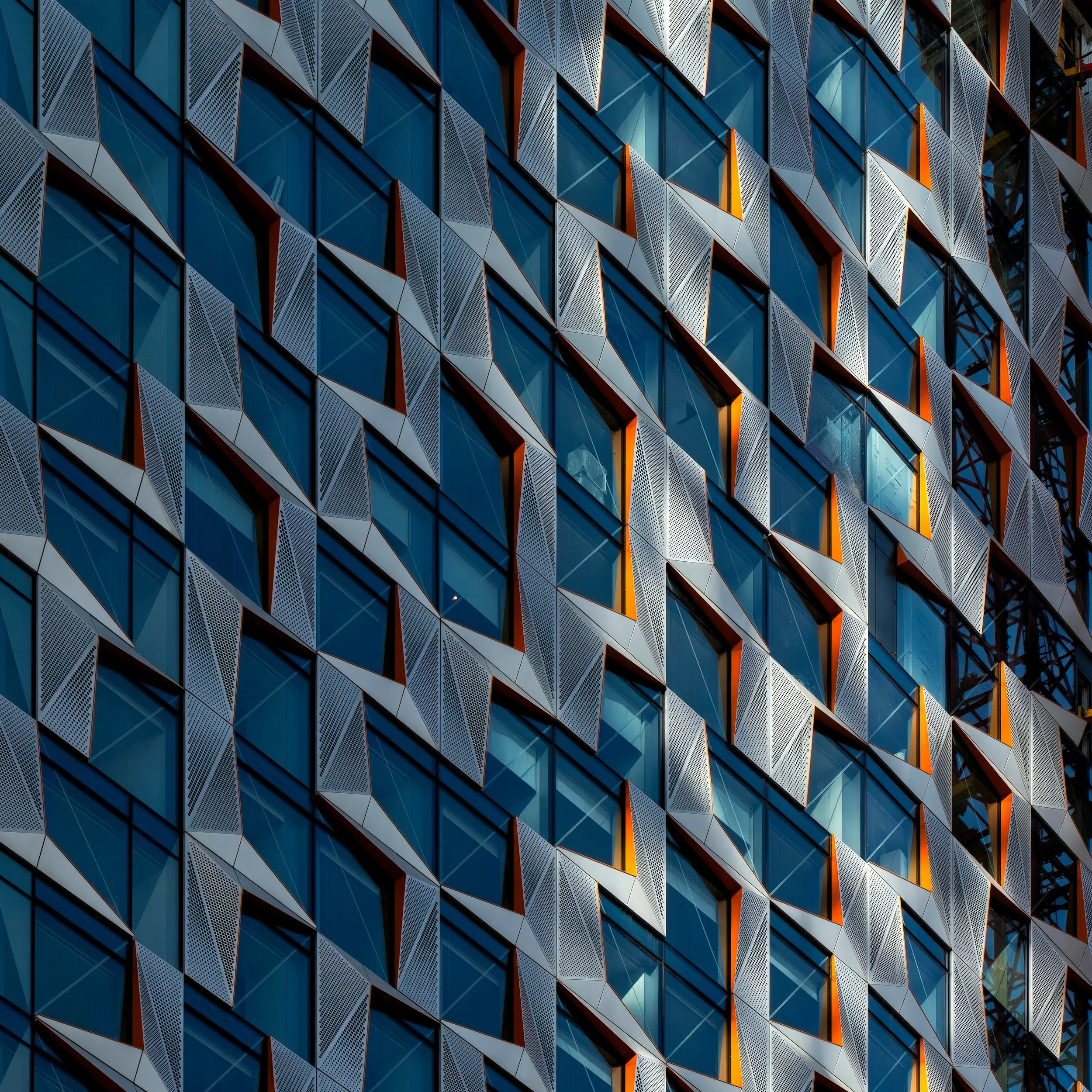 Close-up van een modern gebouw met een gevel van blauwe glazen ramen en metalen panelen met perforaties, afwisselend met oranje accenten.