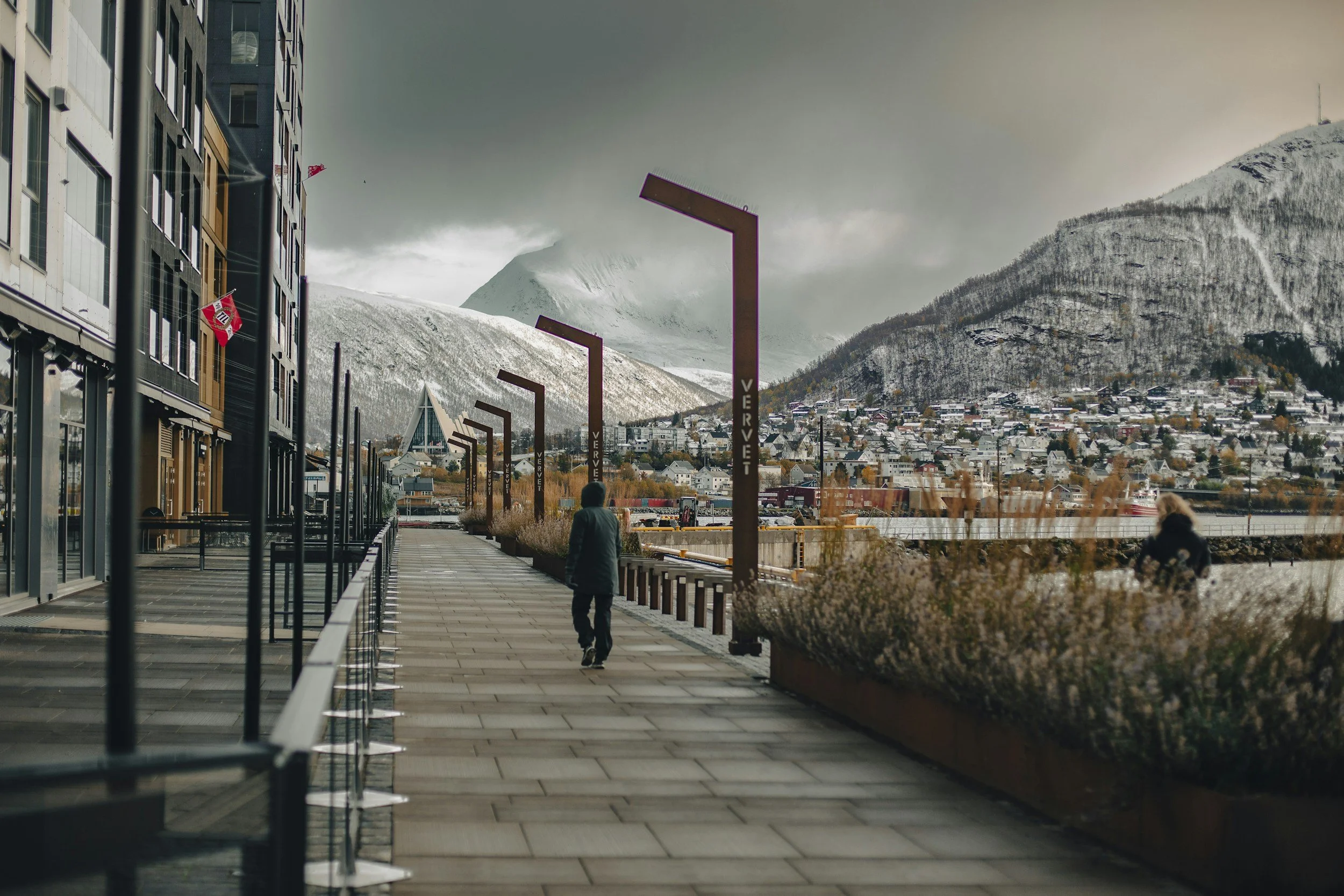 Utsikt over en by med fjell i bakgrunnen, noen personer går langs en promenade, og det er et moderne bygg på venstre side av bildet.