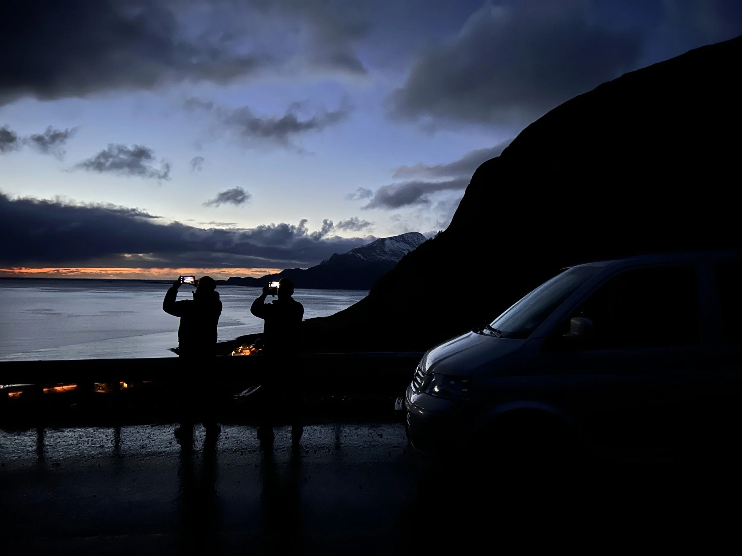 To personer står ved vannet og tar bilder med mobilene sine under en overskyet kveld med fjell i bakgrunn, mens det er flere biler parkert nær stranden.