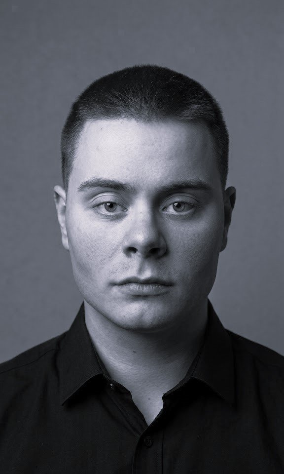 Dødt svart-hvitt portrett av en ung mann med kort hår, iført svart skjorte, mot en grå bakgrunn.