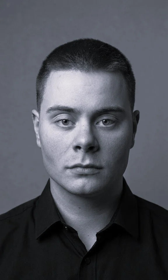 Ung mann med kort, mørkt hår, svart skjorte, ser direkte inn i kamera, i sort-hvitt.