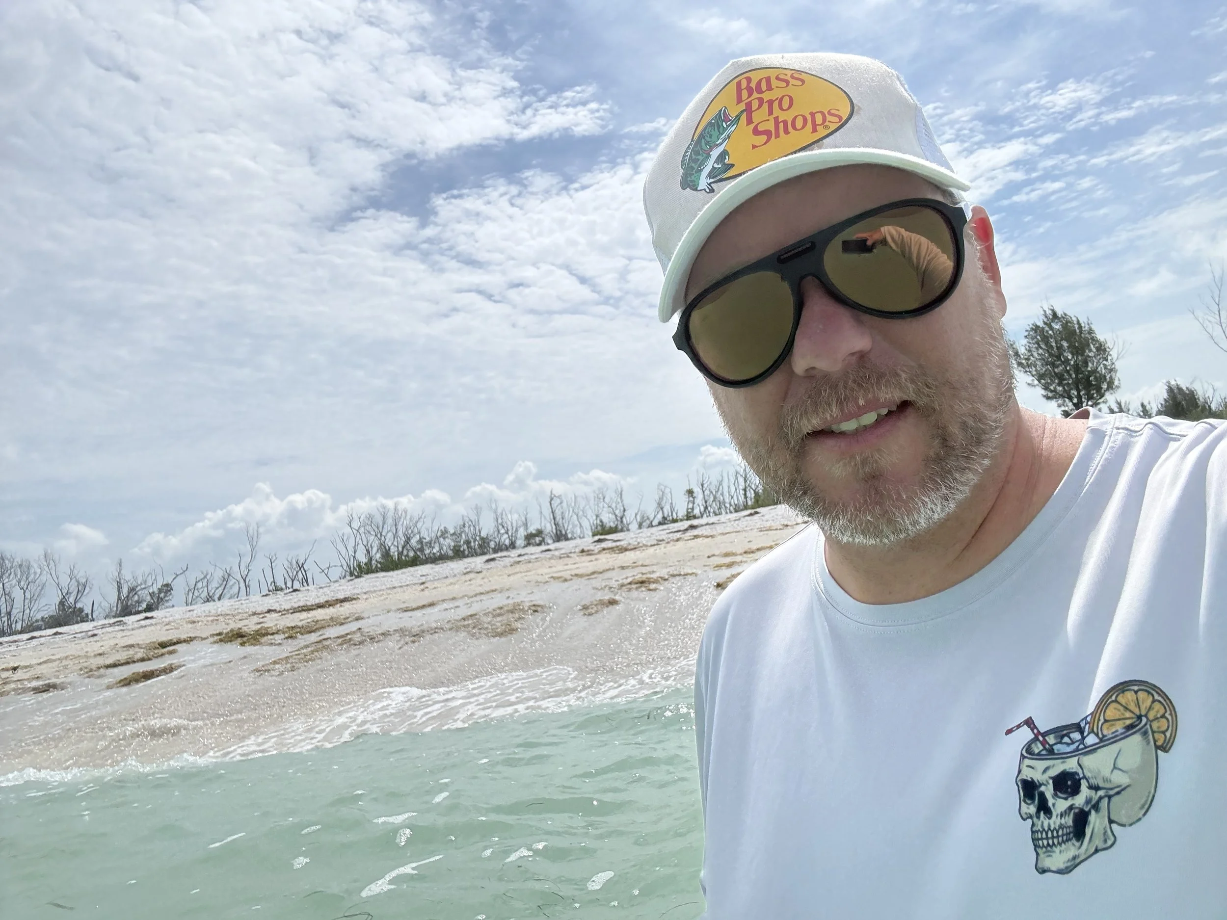 A man with a beard wearing sunglasses, a white hat with a "Bass Pro Shops" logo, and a white T-shirt with a skull and lemon graphic, standing near turquoise water on a sandy beach with a cloudy sky overhead.