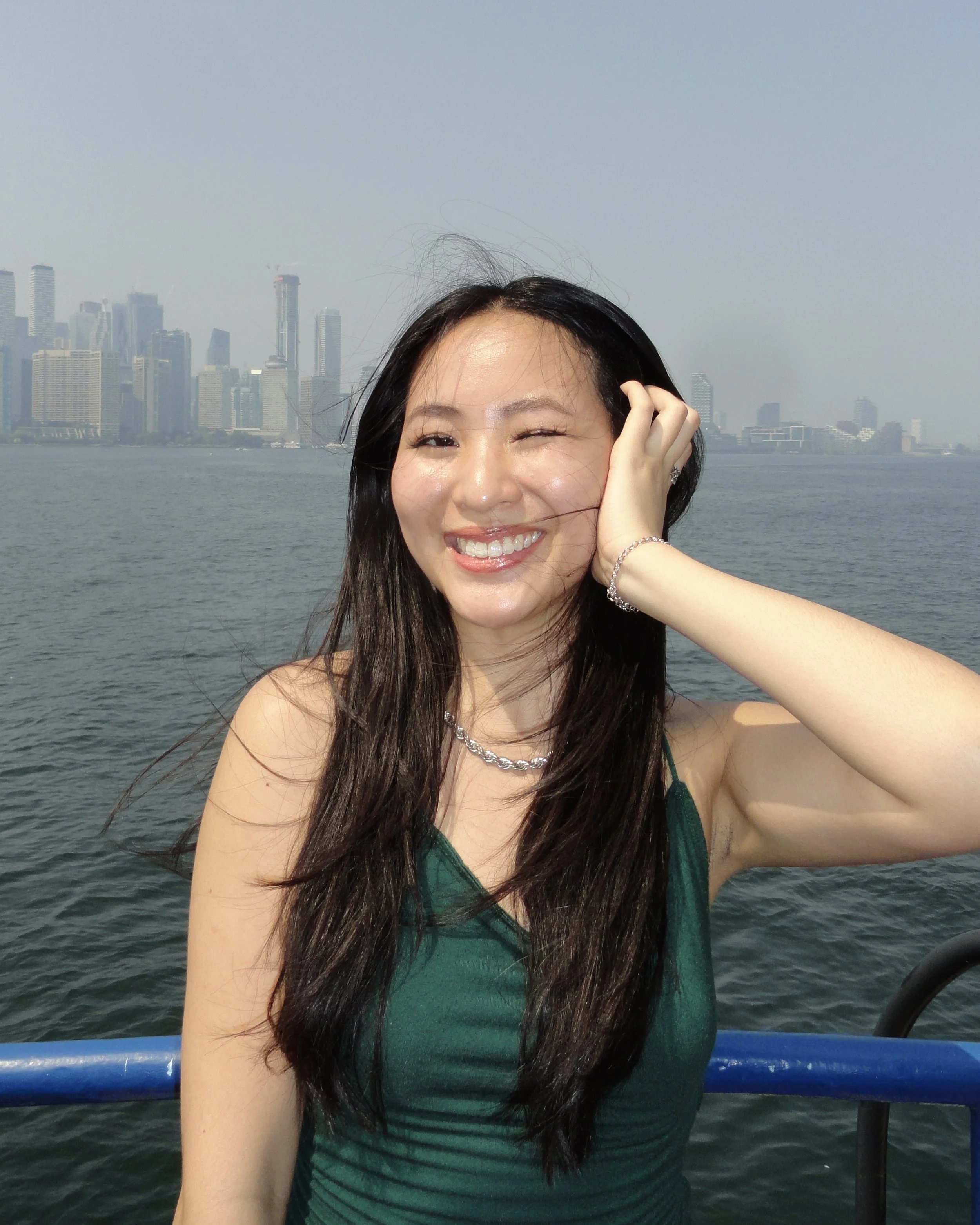 A woman with long dark hair smiling and winking, wearing a green dress, with a city skyline in the background and a body of water in the foreground.
