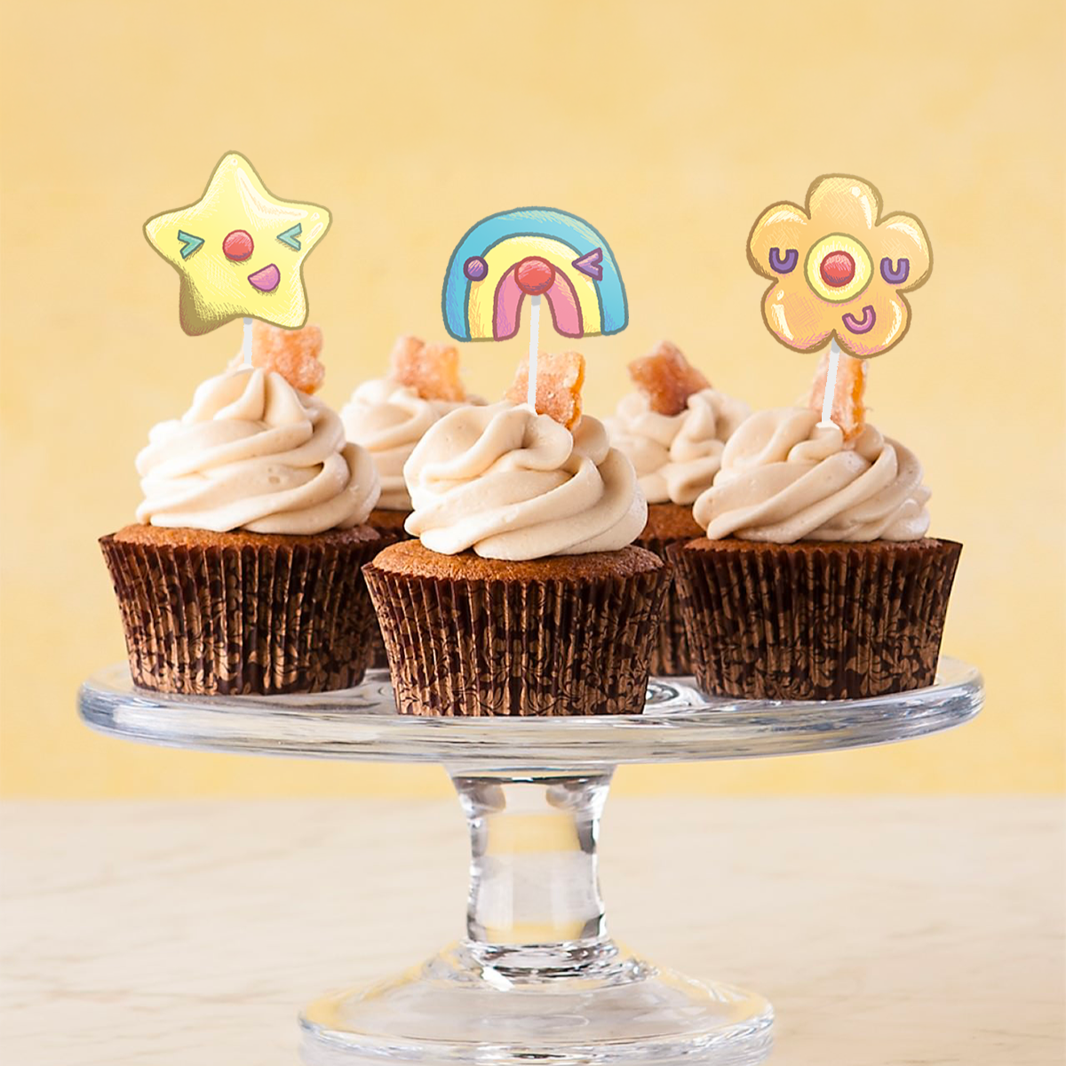 Cupcakes with white frosting topped with colorful cartoon characters on sticks, including a star, rainbow, and flower, on a glass cake stand against a yellow background.