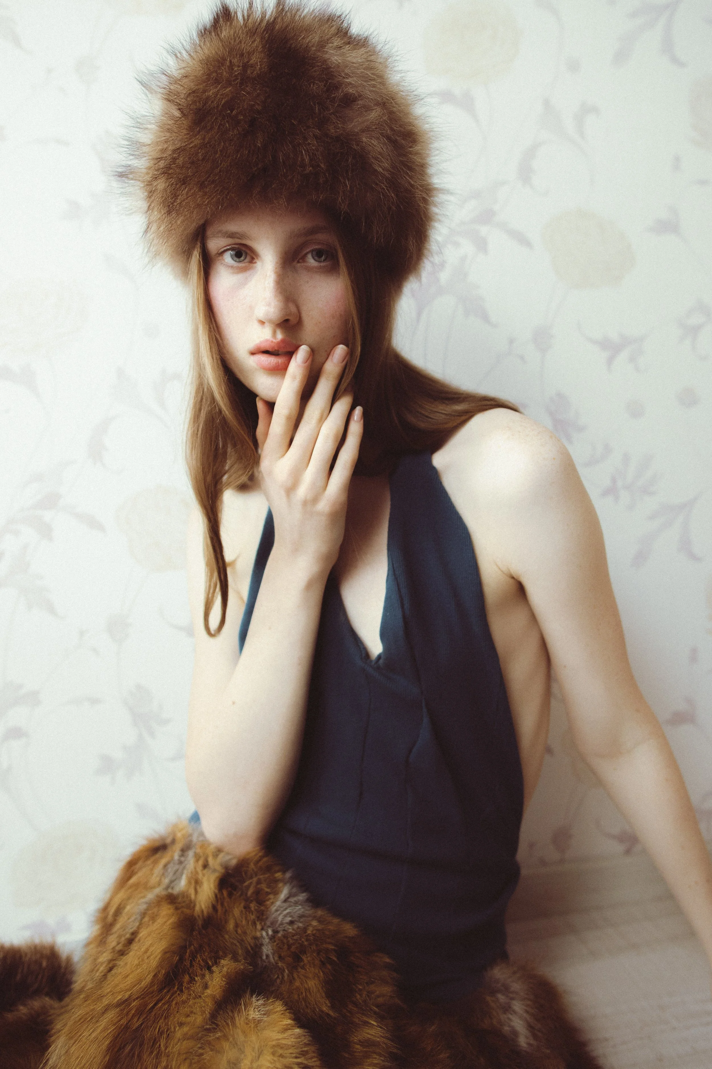 A woman with long light brown hair, wearing a dark blue sleeveless top, is touching her lips with her right hand. She is wearing a large furry brown hat and has a furry animal, possibly a dog or a fox, near her lap. The background features a light-co