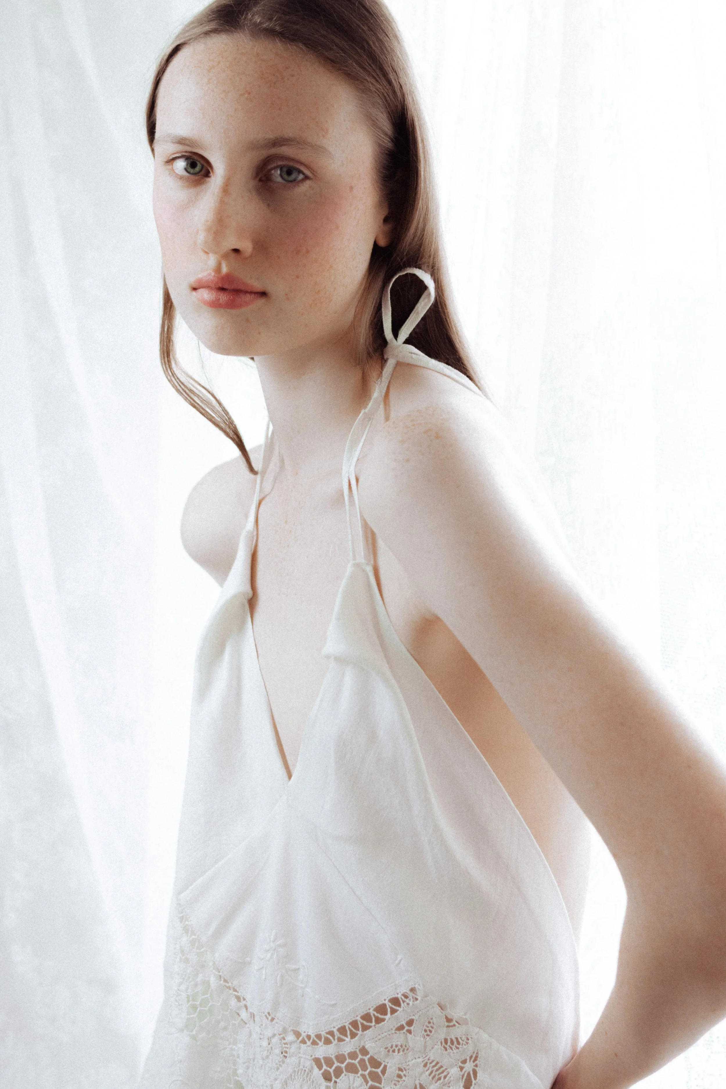 Close-up of a woman with long brown hair, green eyes, and freckles, wearing a white sleeveless top with lace and scalloped edges, standing in front of a sheer white curtain.