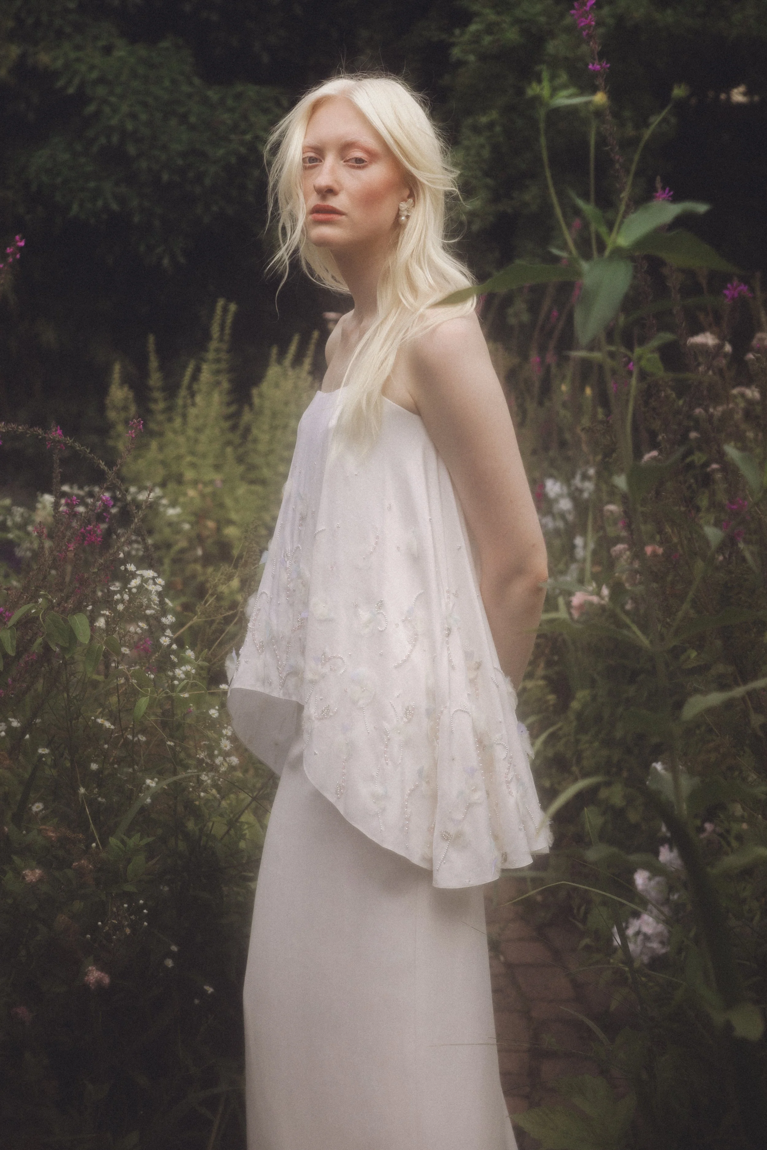 A young woman with long blonde hair standing outdoors among bushes and flowers, wearing a white dress with embroidered details, looking at the camera with a neutral expression.
