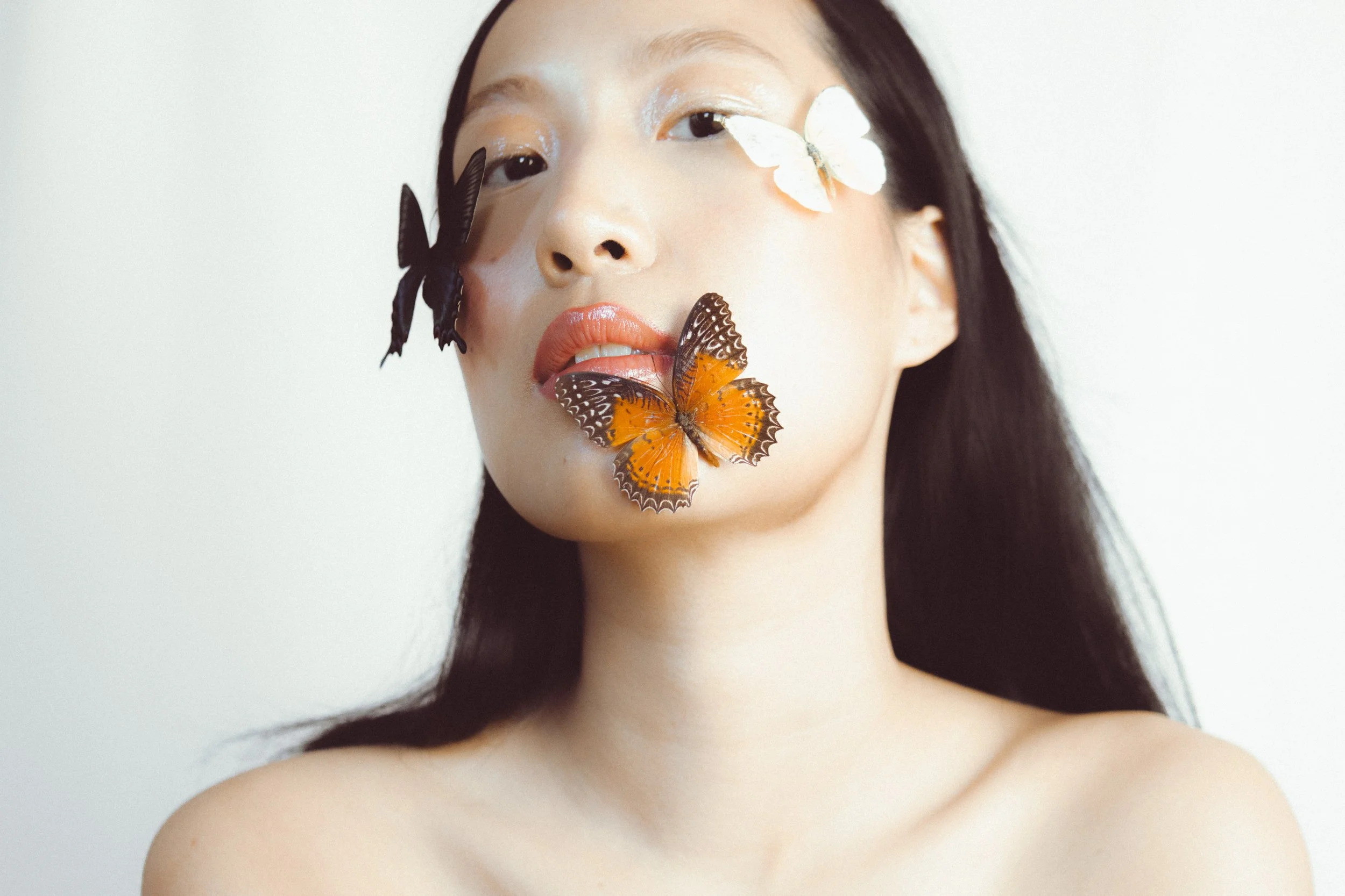 A young woman with long dark hair and light skin, with butterflies on her face and lips, against a plain light background.