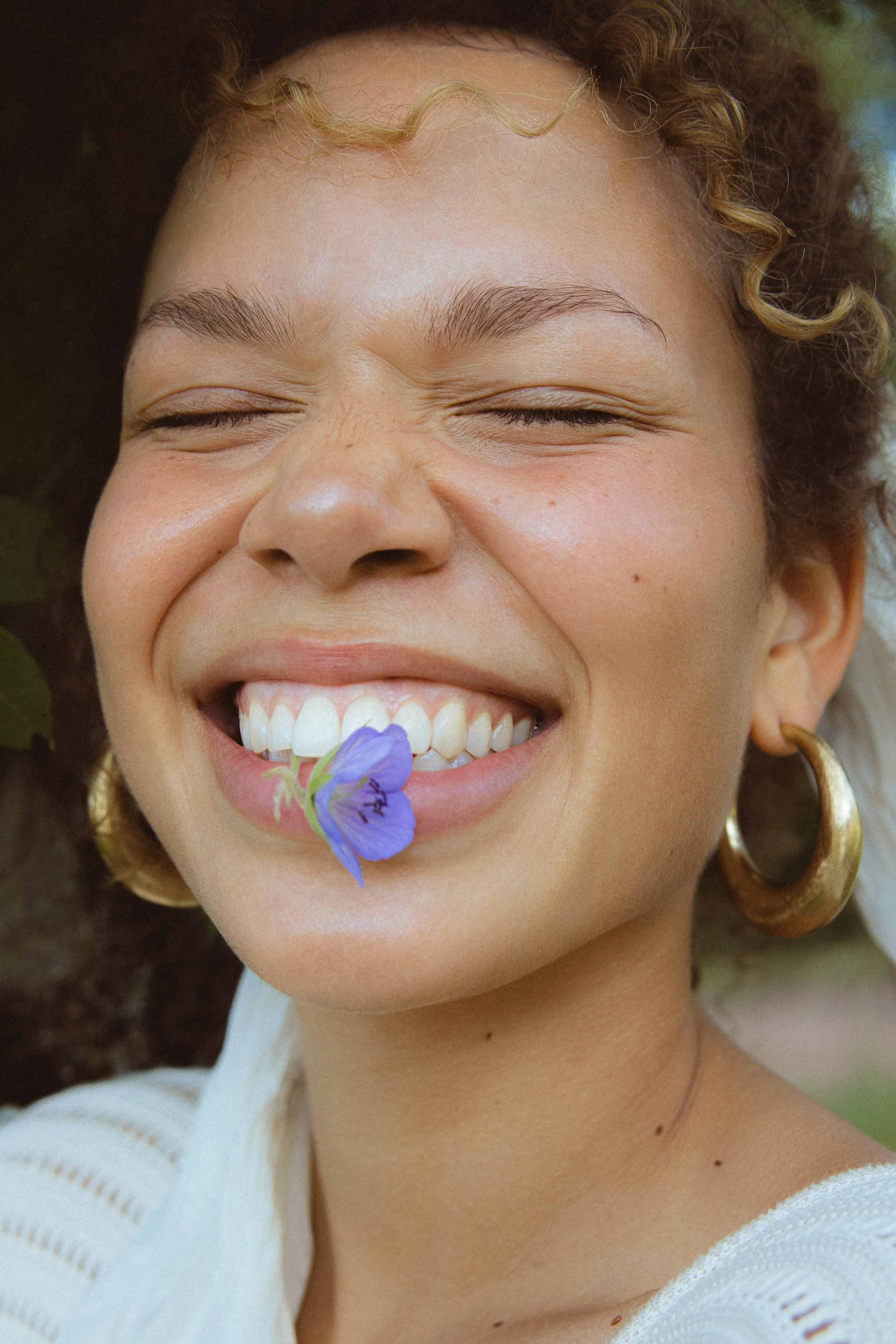 A woman with curly hair and gold hoop earrings smiling with her eyes closed, holding a purple flower near her mouth.