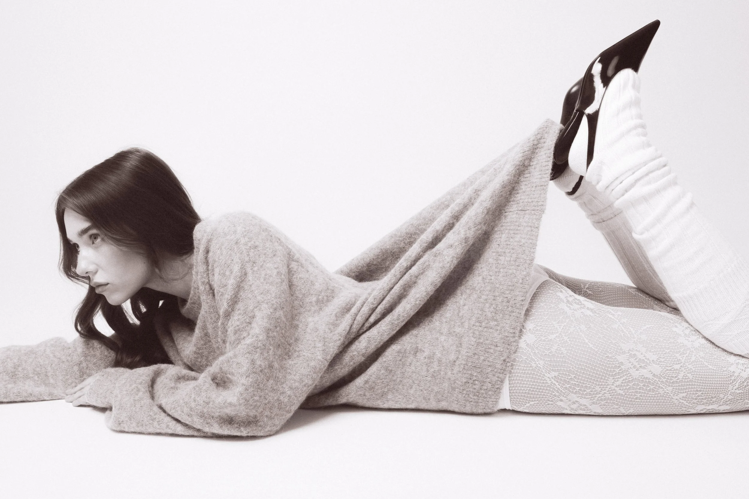 A woman lying on her stomach on a white surface, wearing a large sweater, patterned stockings, and black pointed shoes, with her legs raised and crossed at the ankles.