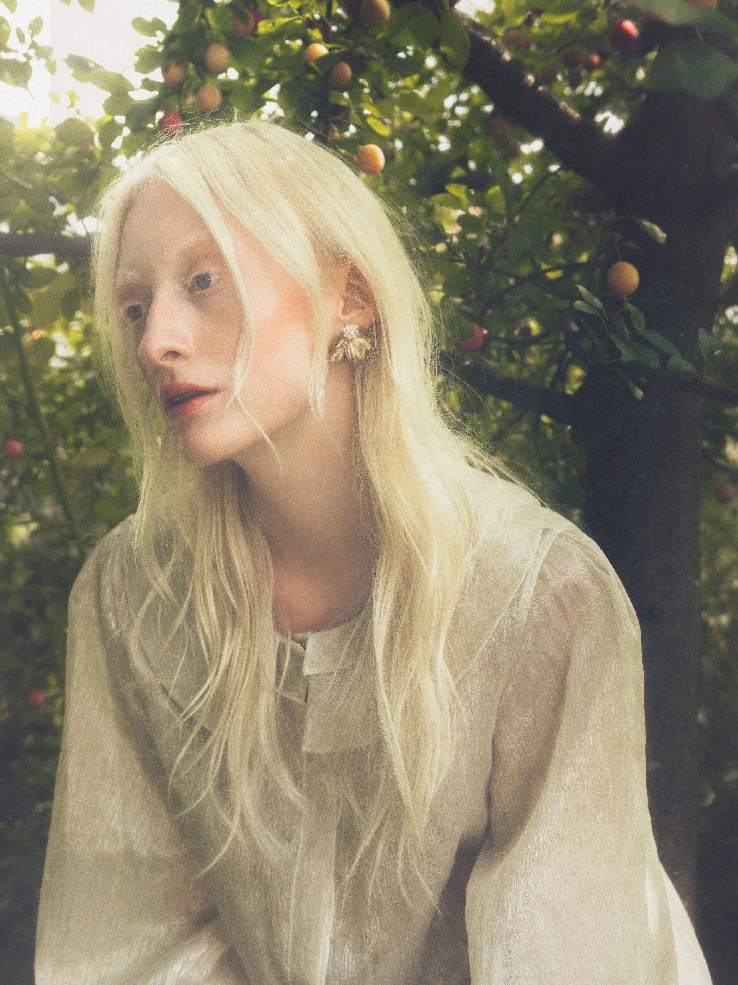 A young woman with long blonde hair and wearing statement earrings, standing near a tree with small round fruits, likely apples or cherries, in a lush outdoor setting.
