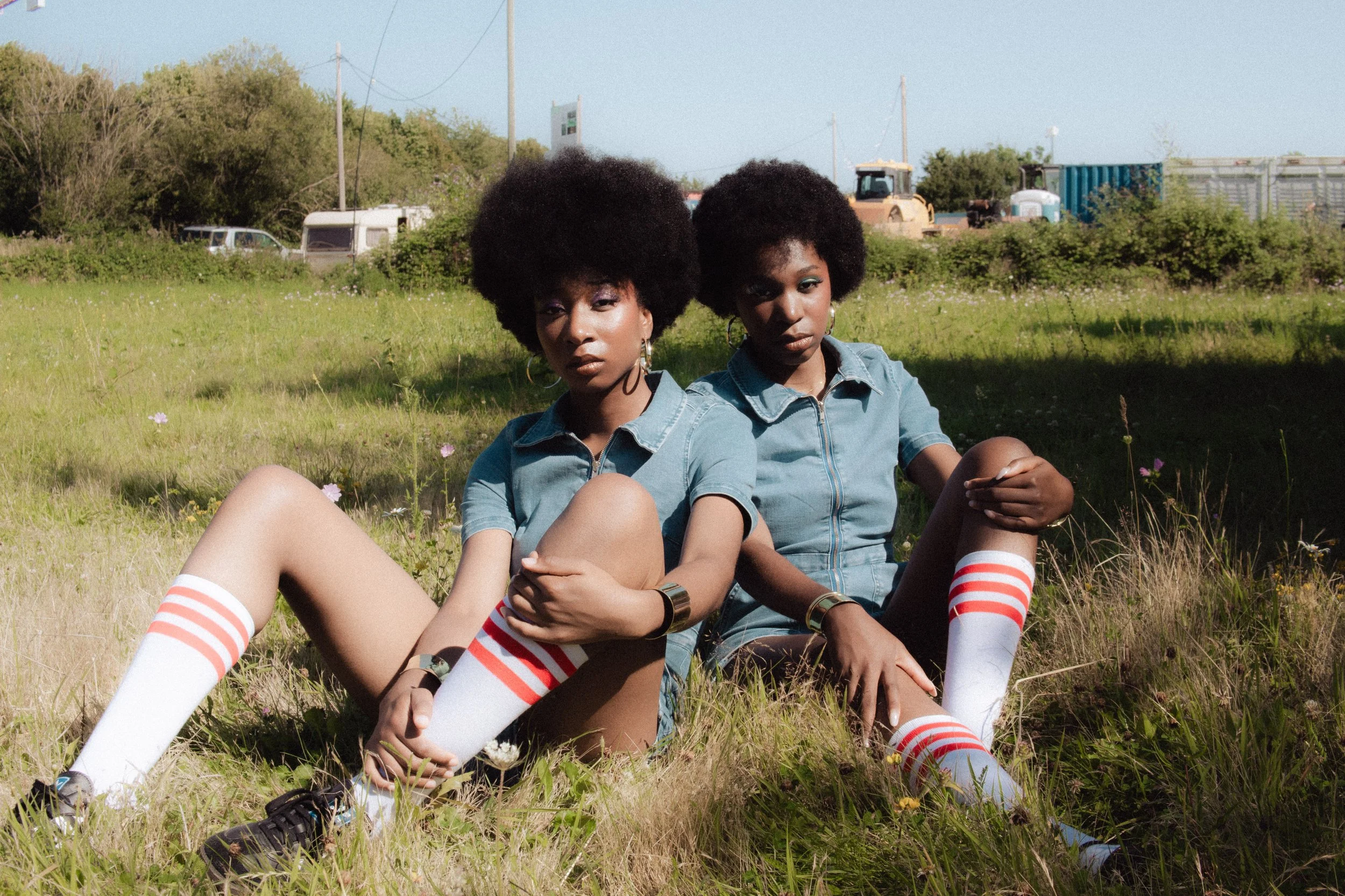 Two women with natural afro hairstyles sitting on grass in a field, wearing denim jackets and striped knee-high socks, with vehicles and trees in the background.