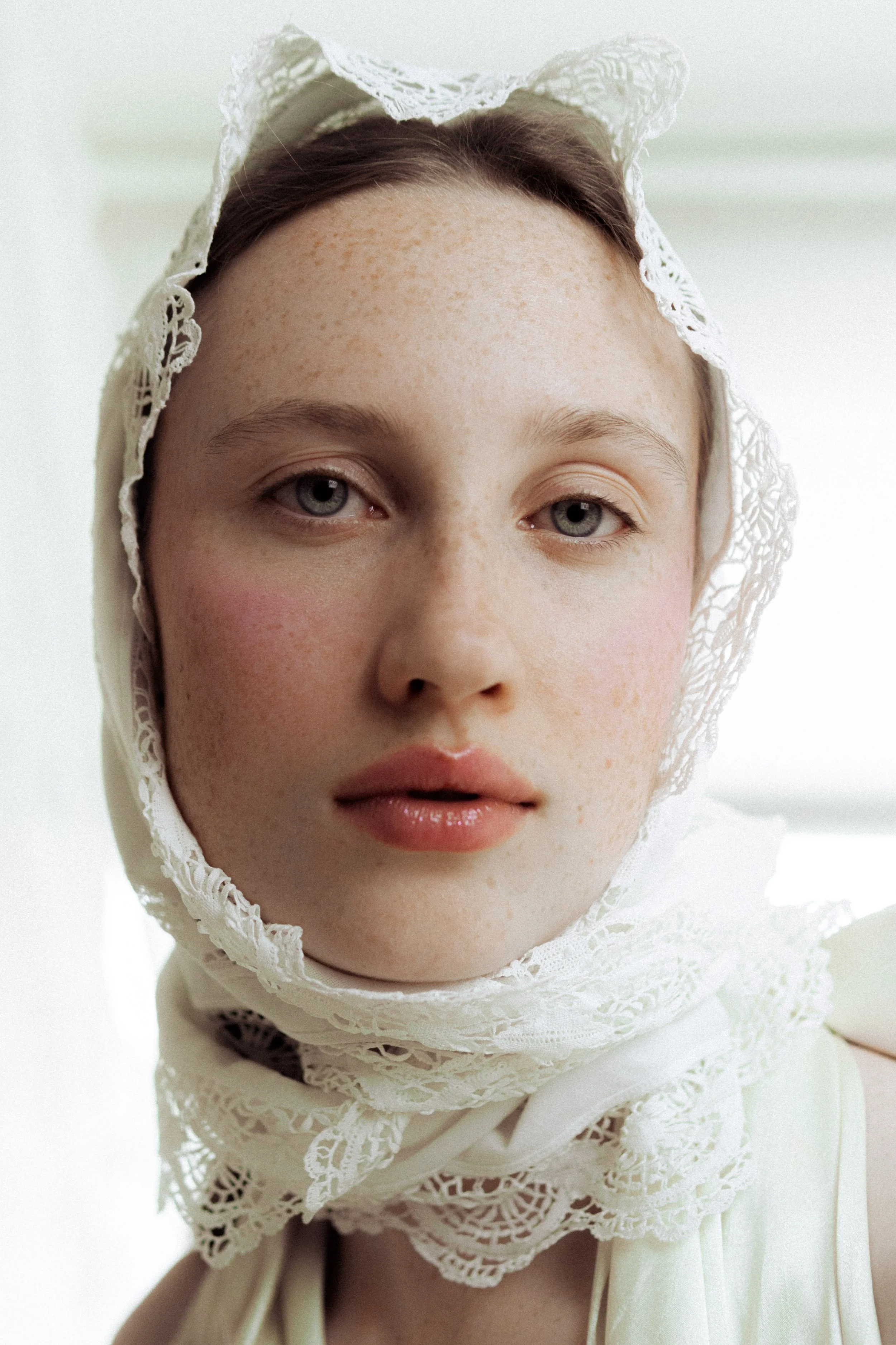 A young woman with blue eyes and freckles wearing a vintage white lace bonnet and matching dress, looking into the camera.