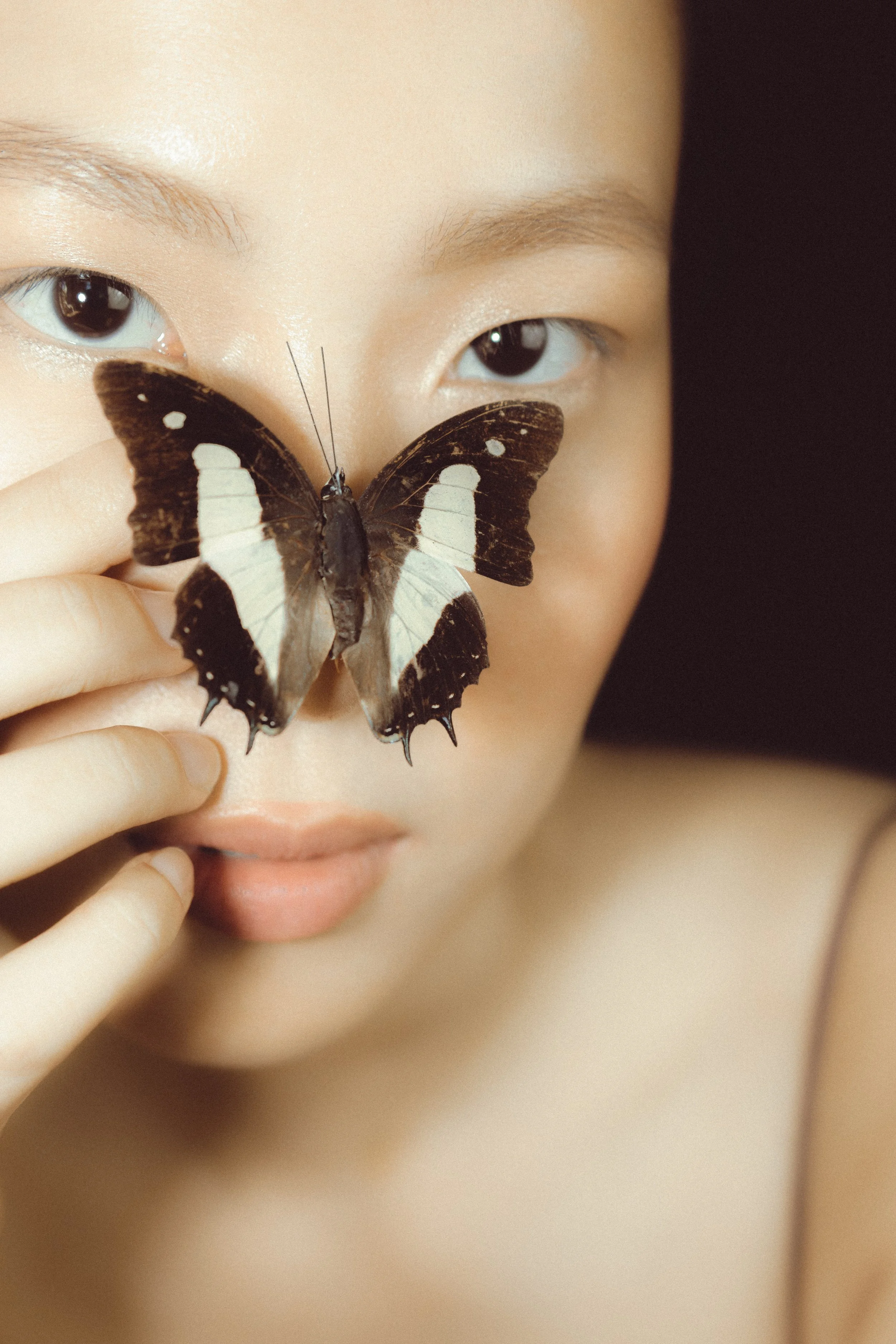 Close-up of a woman's face with a black and white butterfly resting on her nose, holding it gently with her fingers, against a dark background.