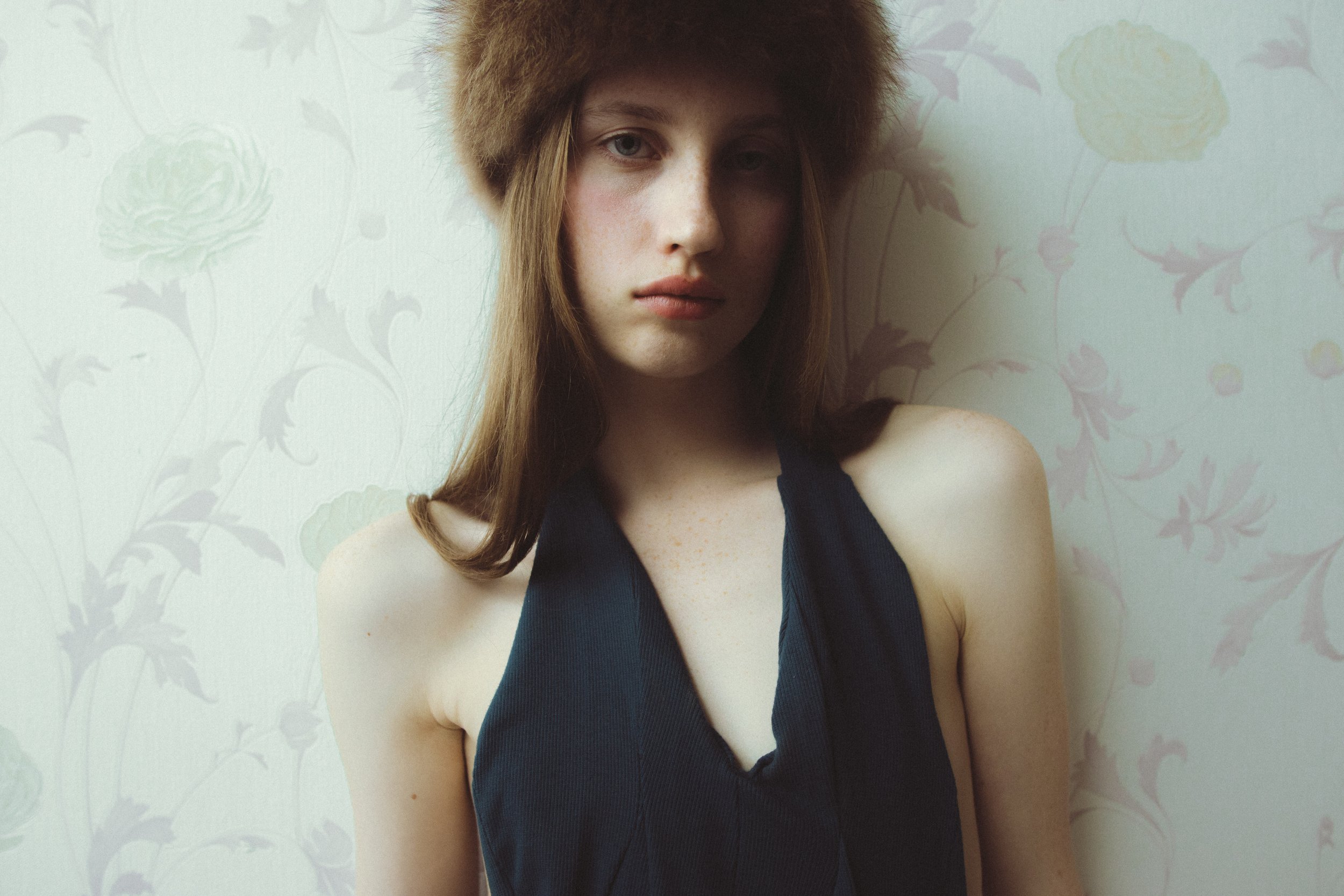 A young woman with long, brown hair wearing a black sleeveless top and a furry hat, standing in front of a light-colored floral wallpaper background.