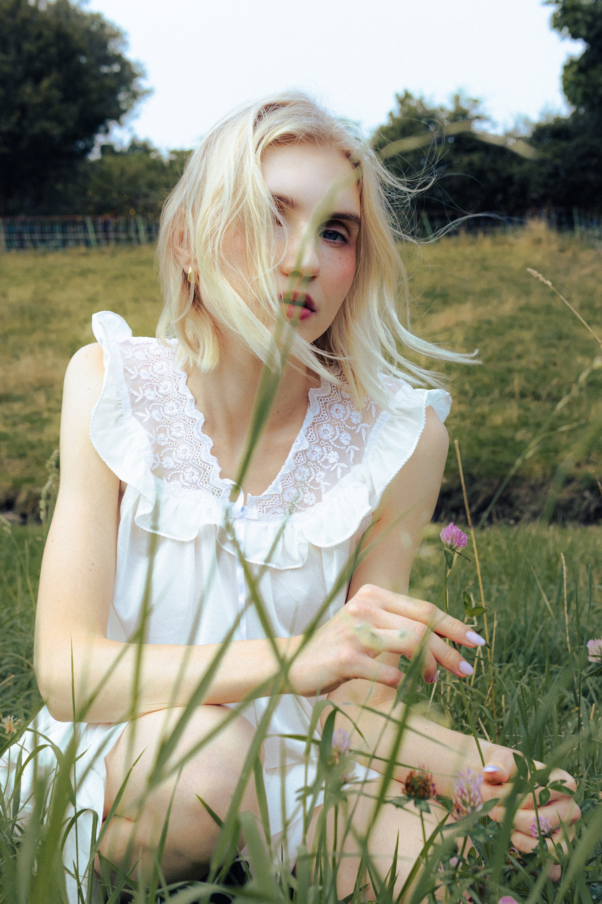 A young woman with blonde hair and blue eyes is sitting outdoors in a grassy field, partially obscured by tall grass and wildflowers. She is wearing a white, sleeveless, embroidered blouse and is holding a flower. The background includes trees and a 