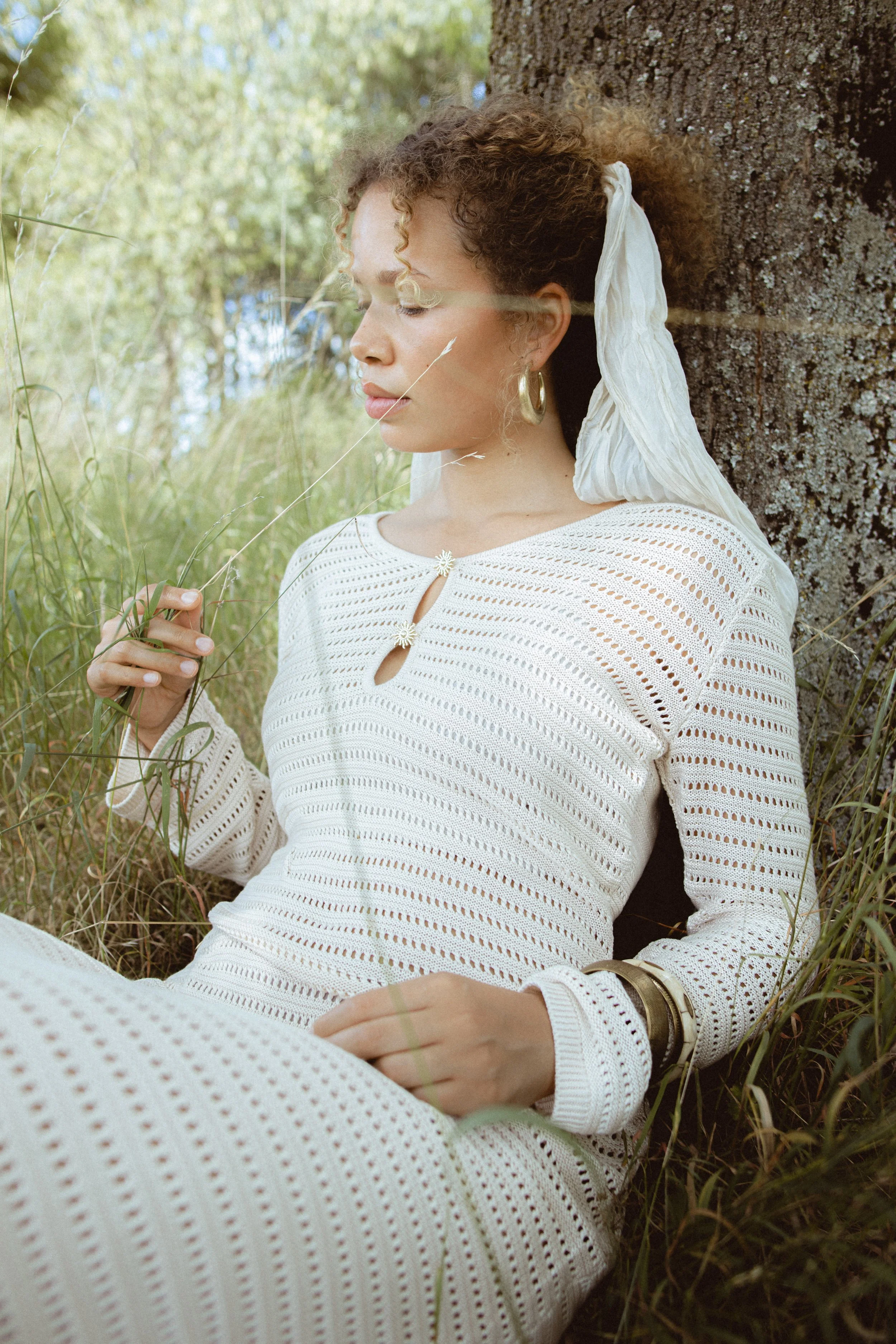 A woman with curly hair wearing gold hoop earrings and a white, knitted dress is sitting outside against a tree trunk. She is gently holding a stalk of grass and appears to be in a peaceful moment with her eyes closed.