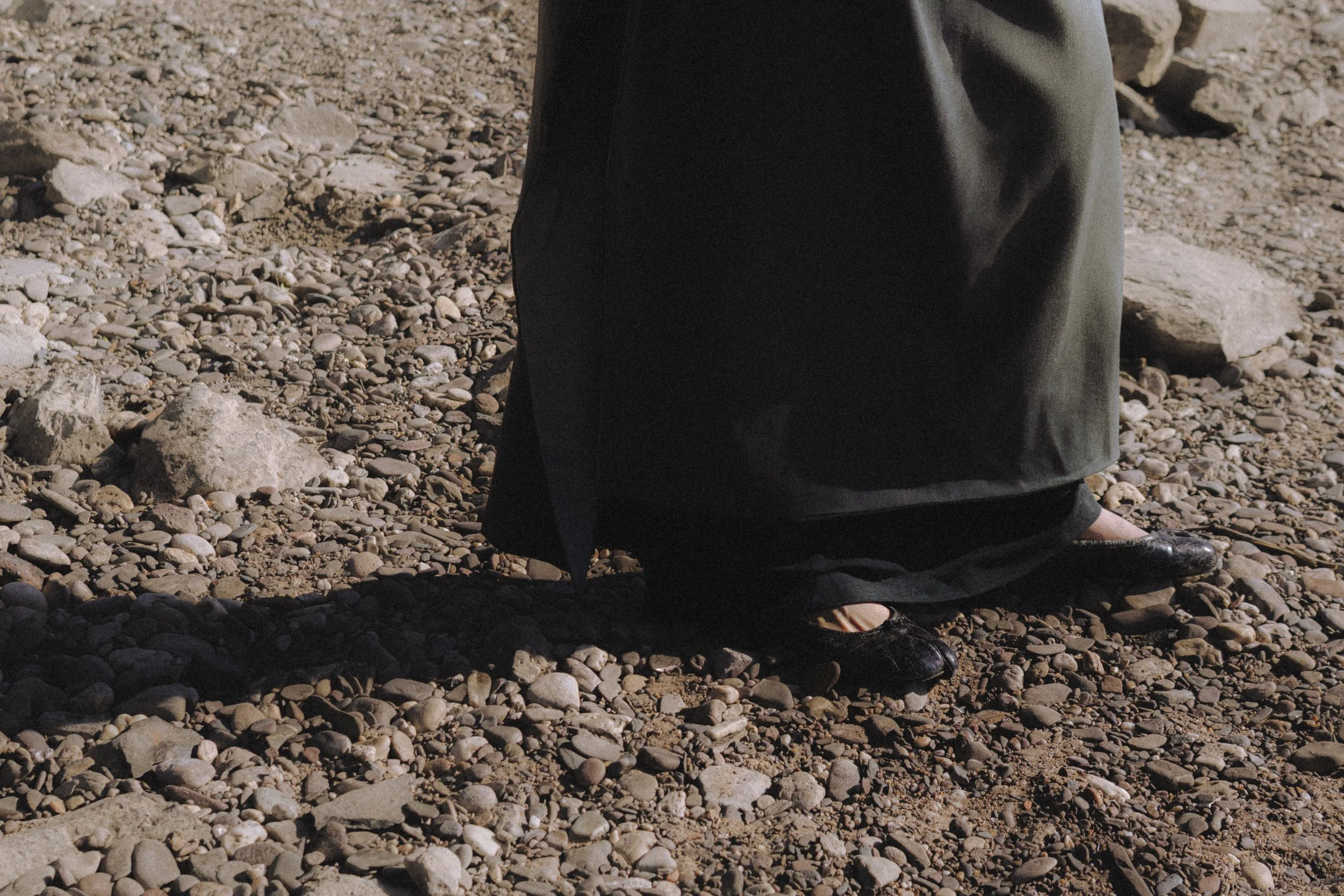 Close-up of a person standing on a rocky beach, wearing black pants and black shoes.