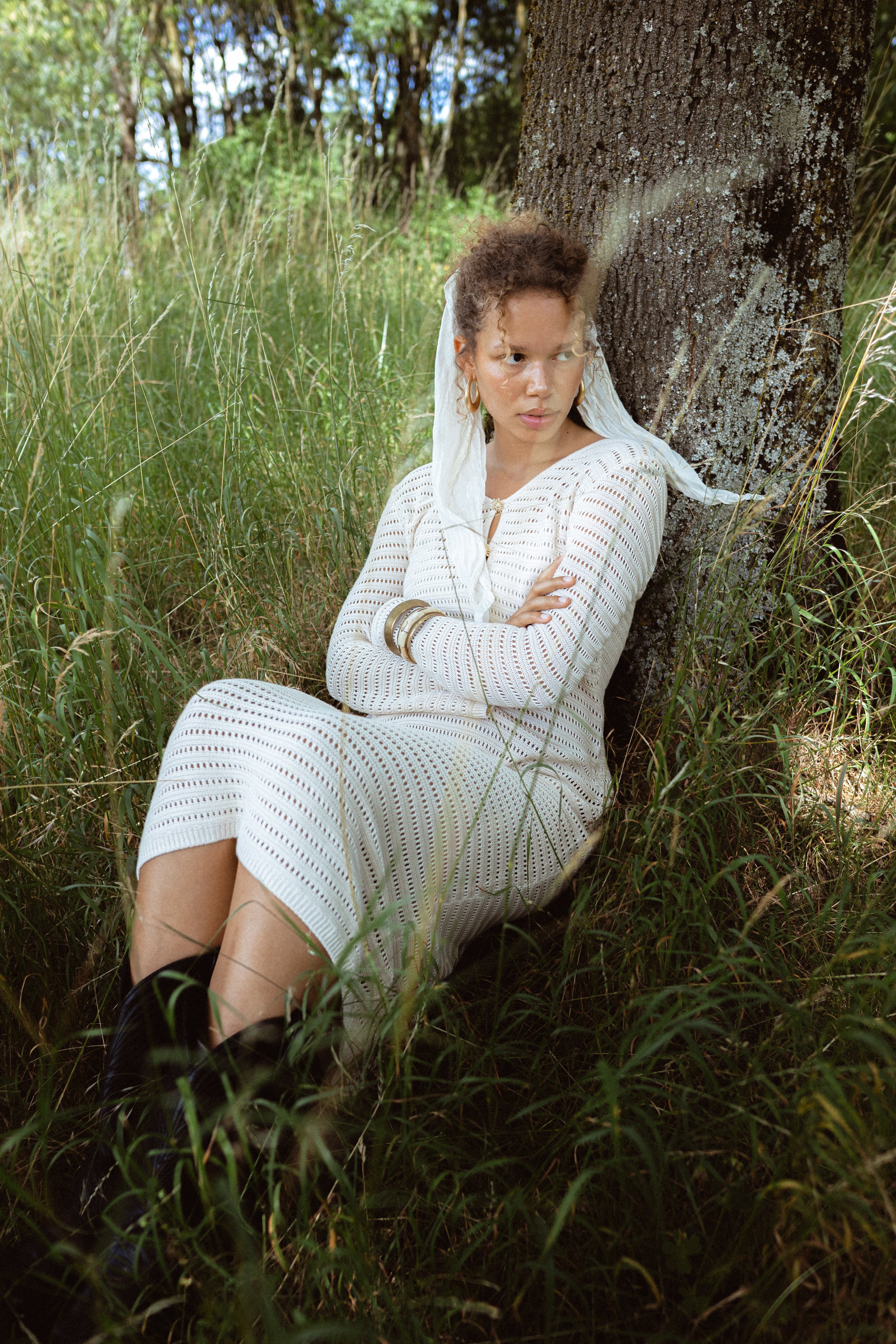 A woman sitting against a tree in a grassy area surrounded by trees, wearing a white crochet dress and gold jewelry.