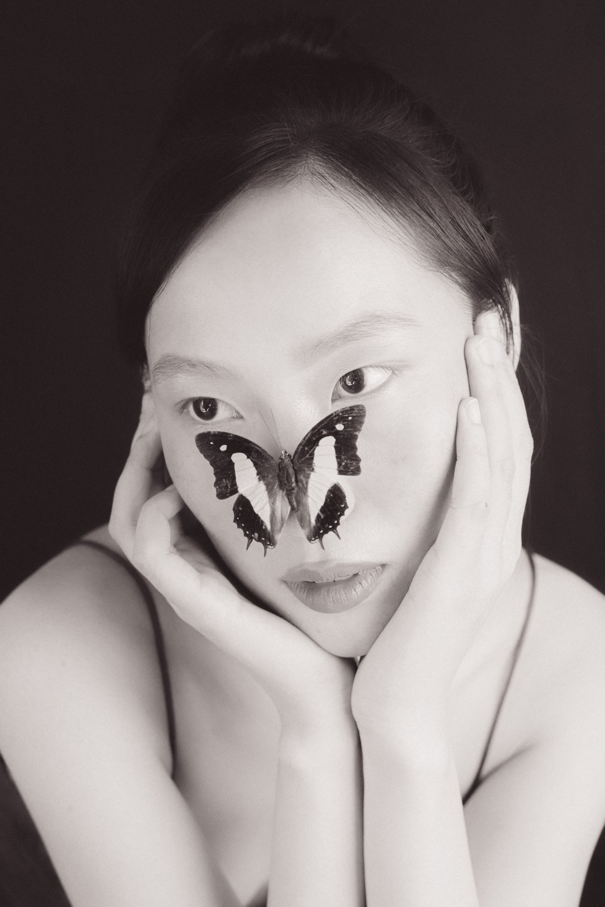 A black and white photo of a woman with a butterfly covering her nose. She is holding her face with both hands, and her expression is neutral. The background is dark, and her hair is styled back.