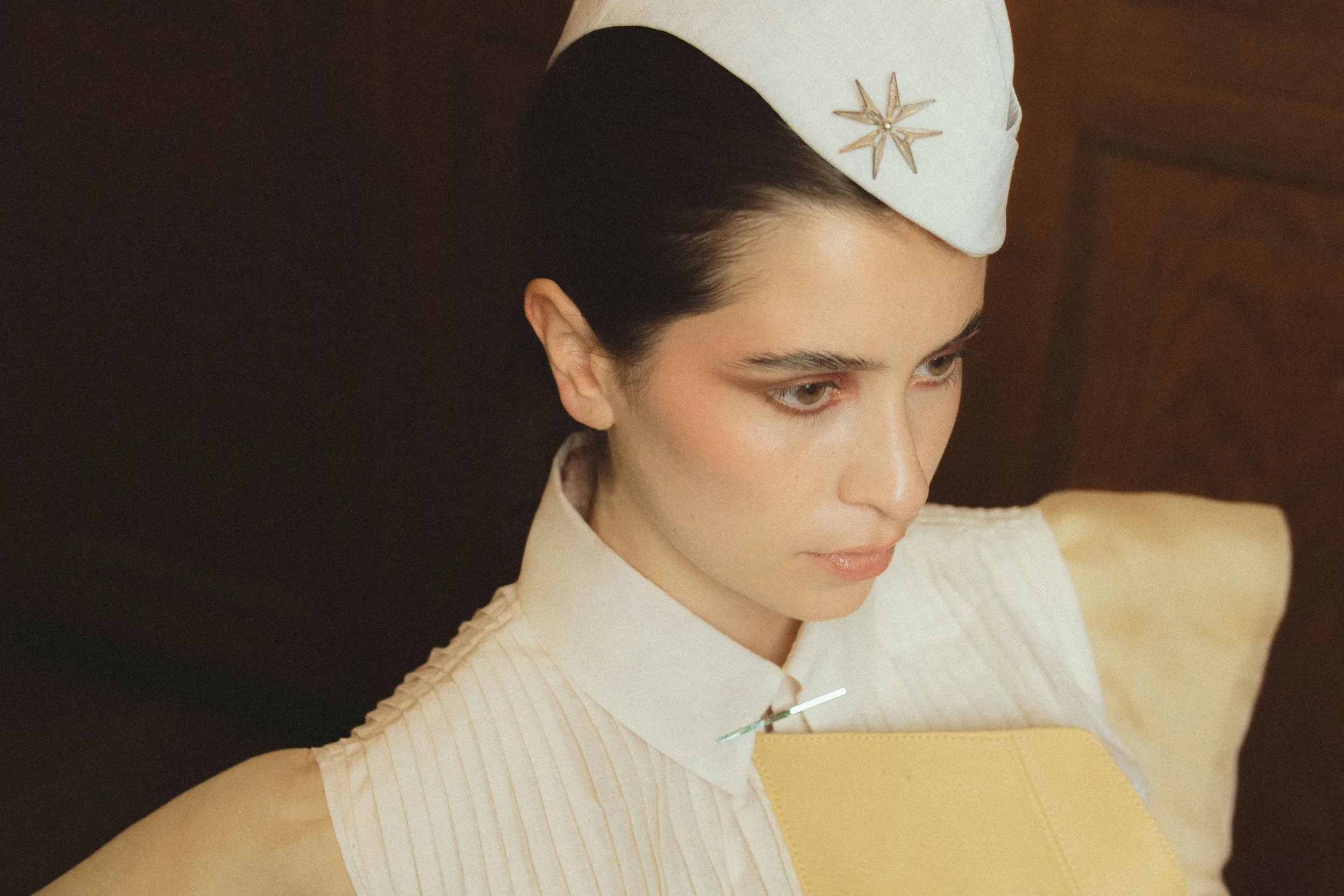 Close-up of a woman with dark hair wearing a white nurse's cap with a star emblem, a white and yellow uniform, and a syringe in her hand, against a dark background.
