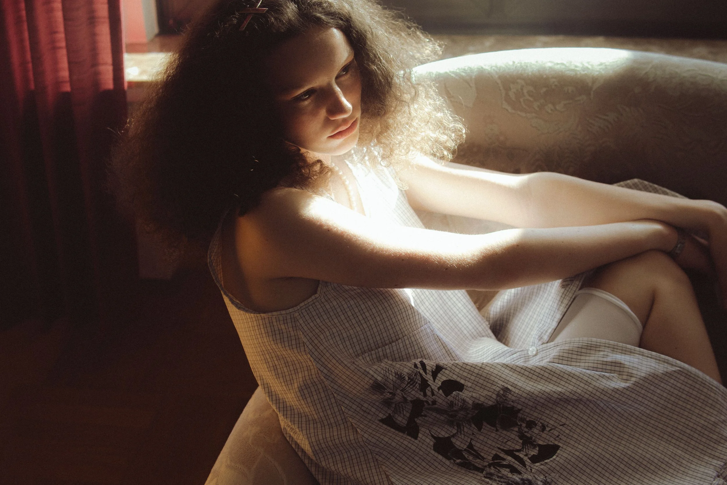 A woman with curly hair sitting by a window with sunlight shining on her face, wearing a checkered dress with black floral print, sitting on a chair with her arms resting on her knees.