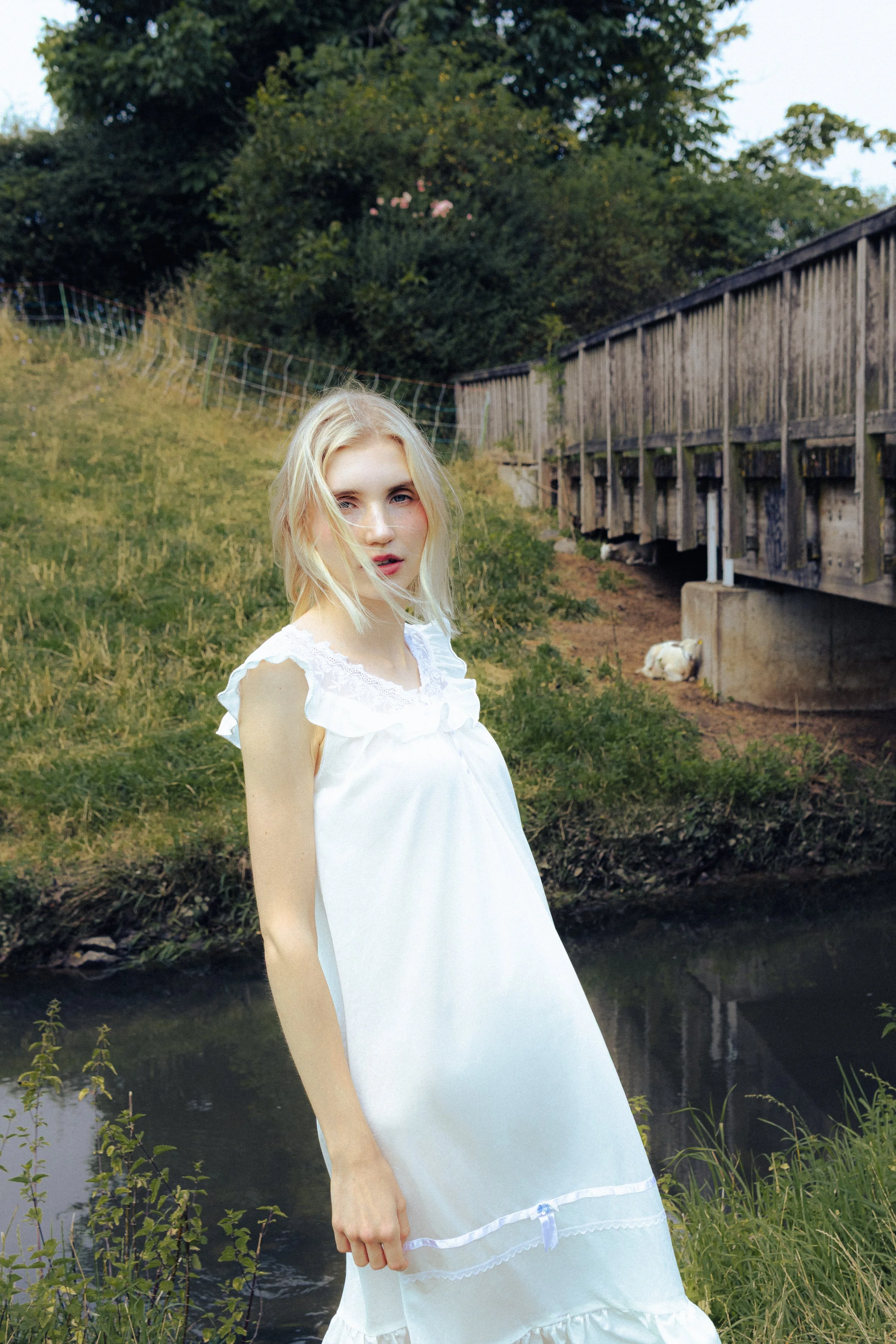 A young woman with blonde hair in a white dress standing near a creek with a wooden bridge and trees in the background.