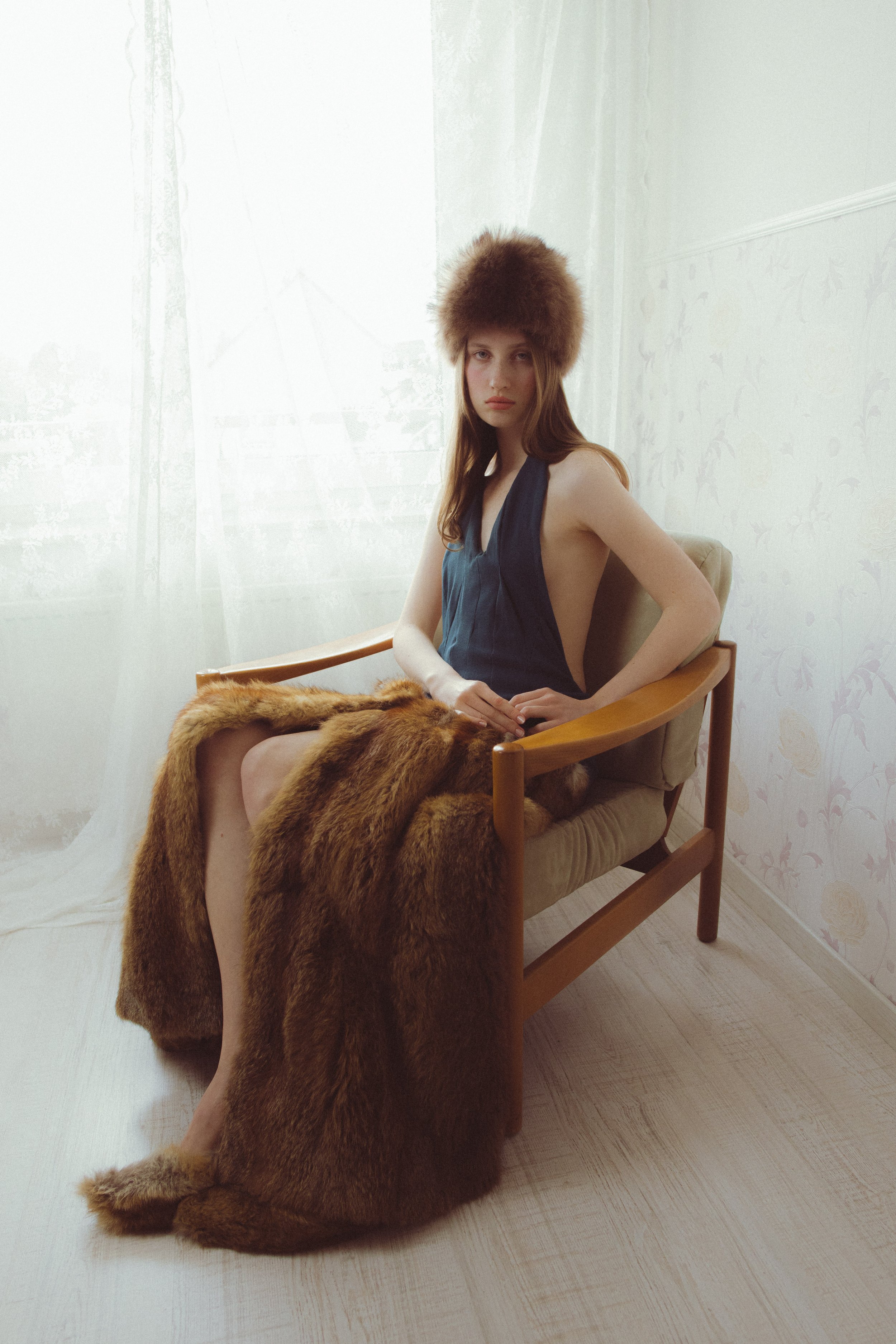A woman with long brown hair sits on a wooden chair, wearing a dark sleeveless top and a large fur hat. There is a fur blanket draped over her lap, and the background includes light-colored wallpaper and semi-sheer curtains with natural light coming 