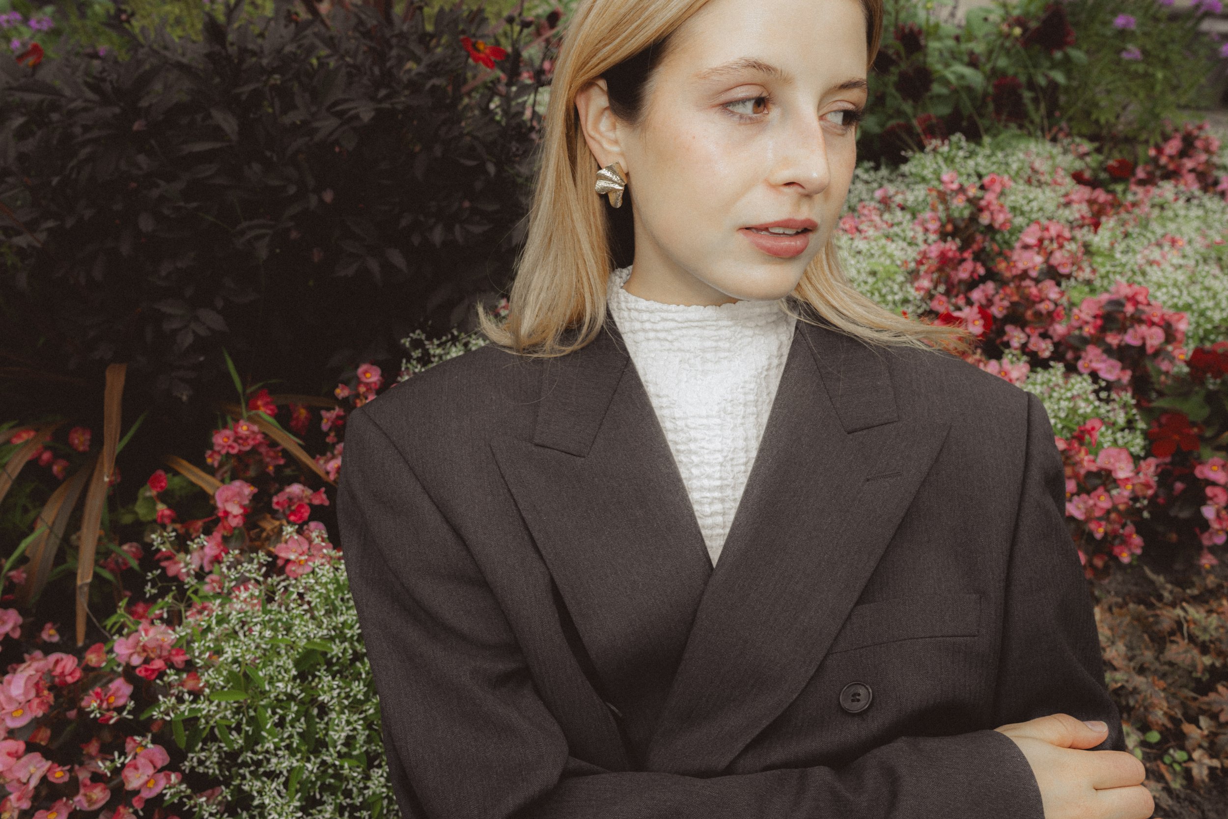 A woman wearing a dark blazer and a white textured turtleneck, standing outdoors in front of pink and white flowers, with her arms crossed and looking to the right.