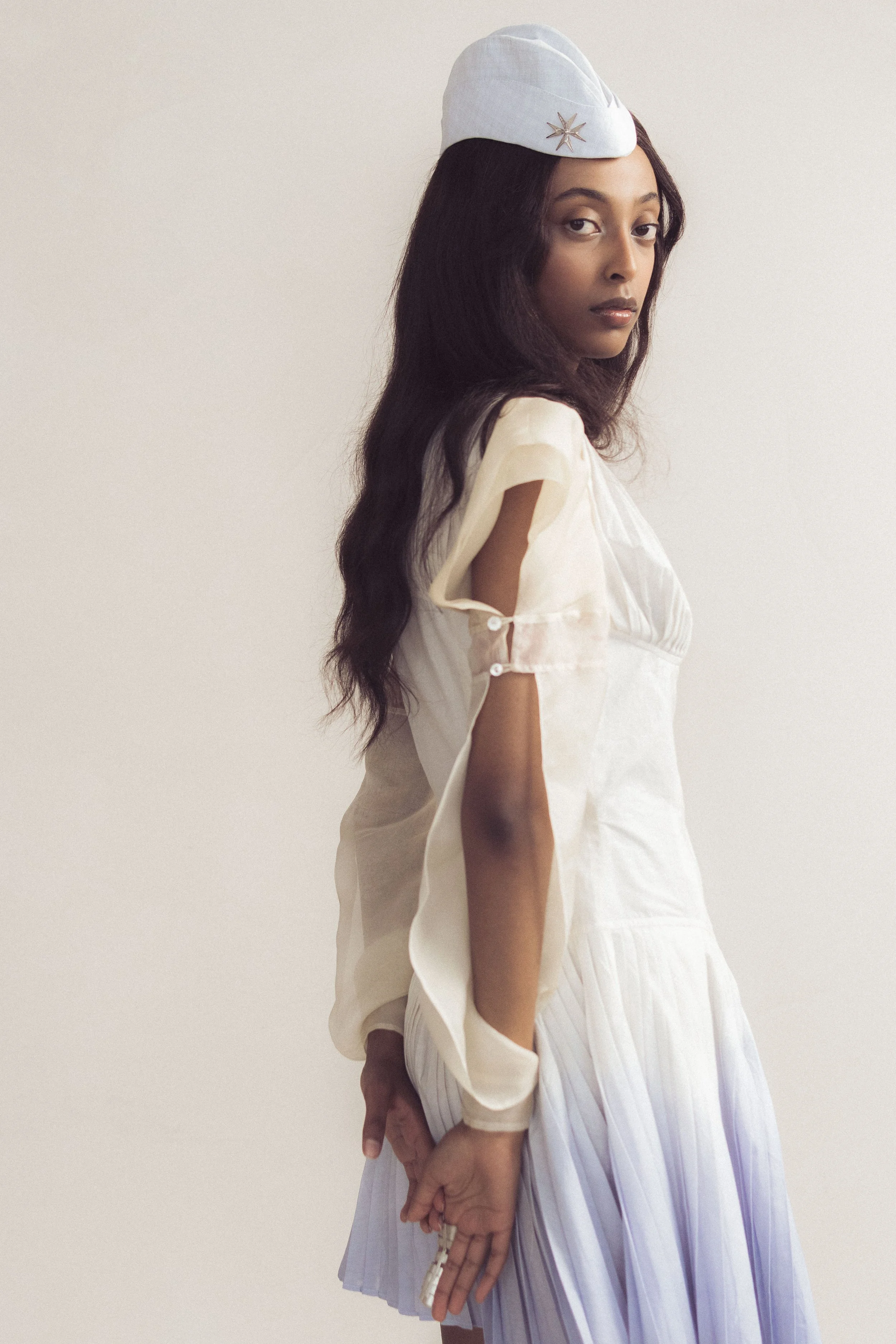 A woman with long dark hair wearing a white hat with a star detail, a light-colored top with shoulder cutouts, and a pleated white skirt, standing against a plain light background.