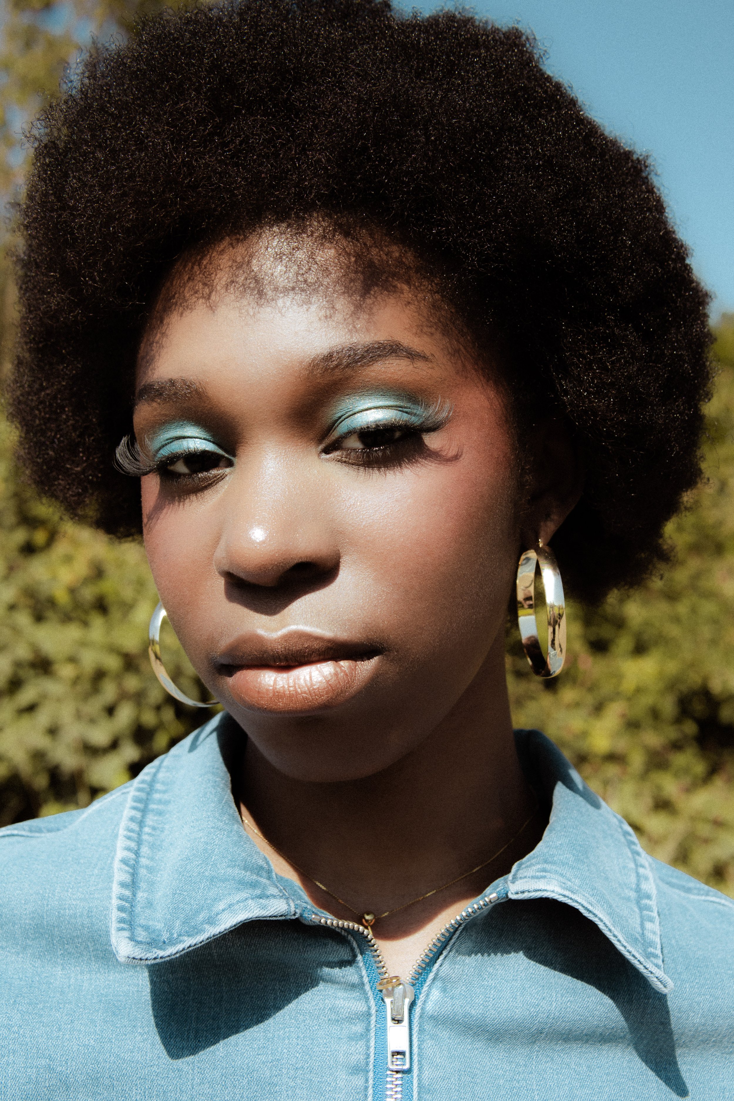 Close-up of a woman with natural curly hair and blue eye makeup, wearing large hoop earrings and a light blue denim jacket outdoors.
