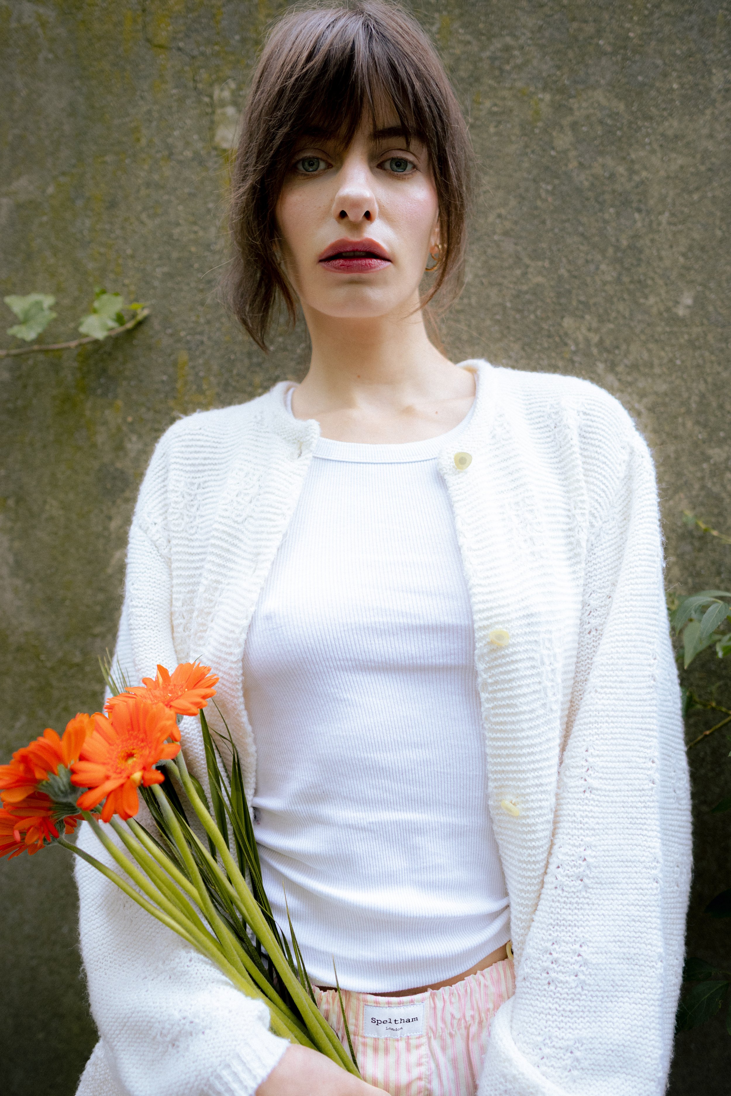 A woman with short brown hair and blue eyes holding a bouquet of orange flowers, standing in front of a concrete wall.