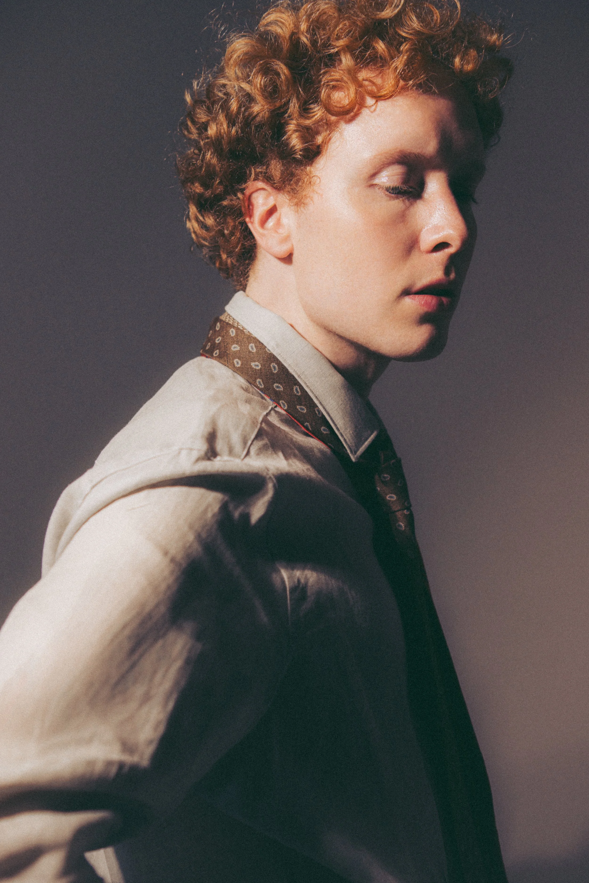 Close-up of a woman with curly red hair, wearing a beige jacket and a patterned shirt, with her eyes closed against a dark background.