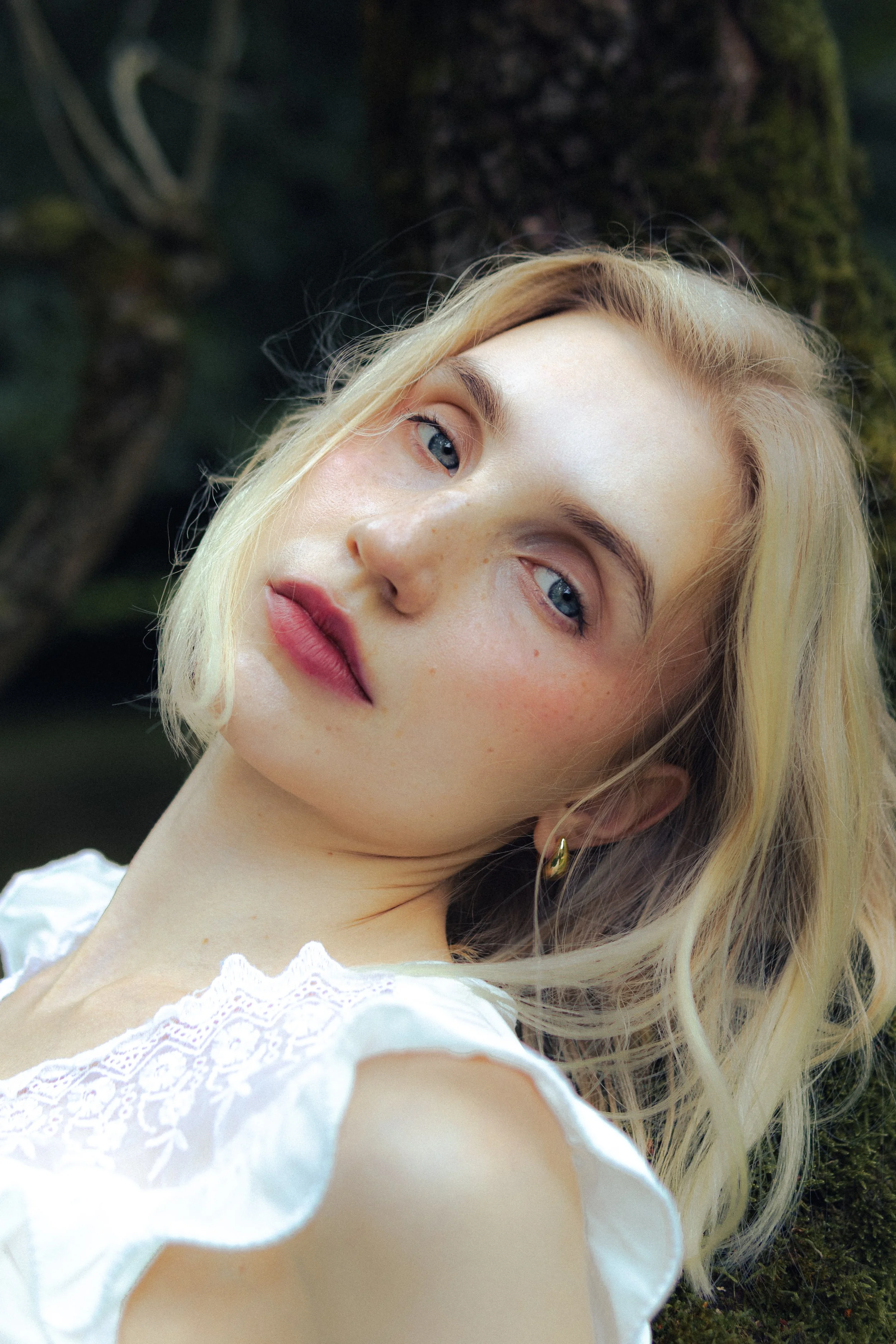 Close-up of a woman with blonde hair and blue eyes, wearing white lace clothing, standing outdoors with greenery in the background.