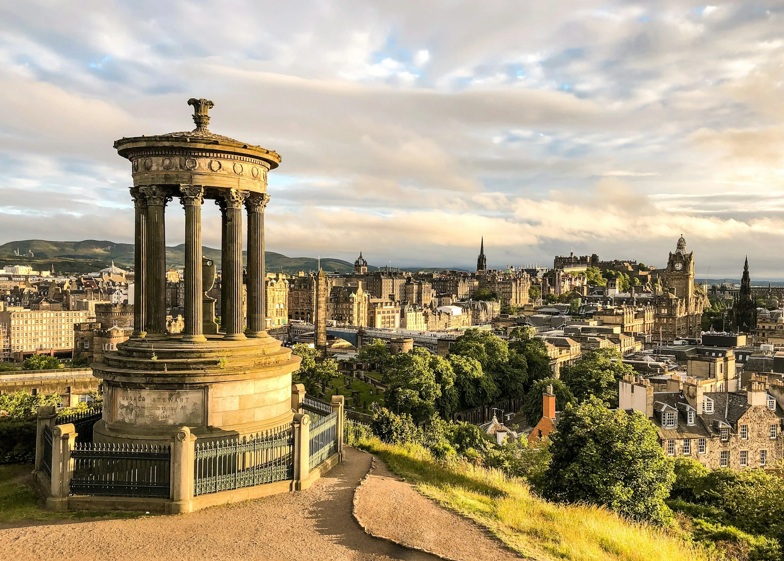 A historic monument with columns and a capstone overlooking the Edinburgh cityscape with old buildings and landmarks, set against a partly cloudy sky during sunrise.