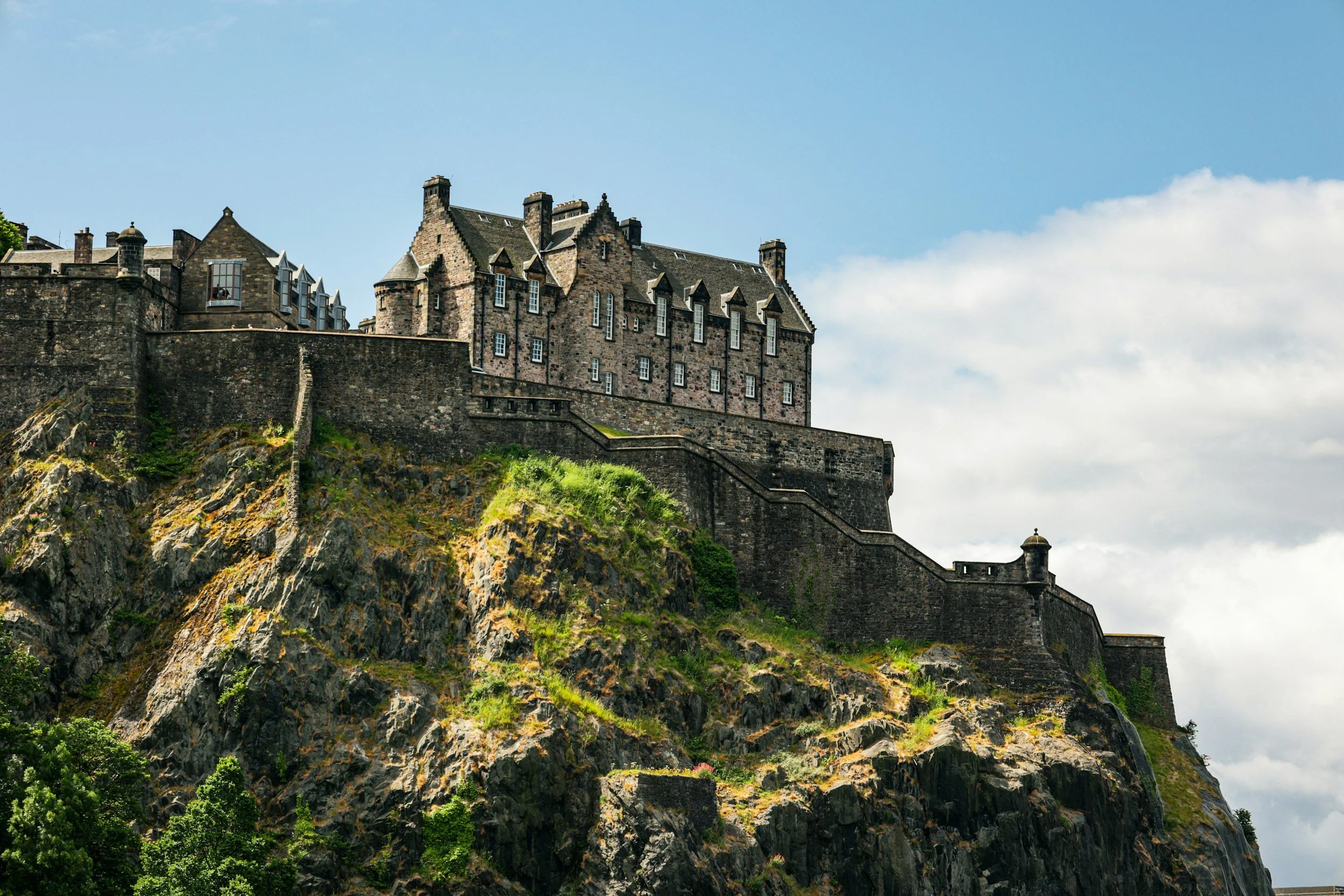 An image of Edinburgh castle situated on top of a rocky hill with a stone wall surrounding it, under a partly cloudy sky.