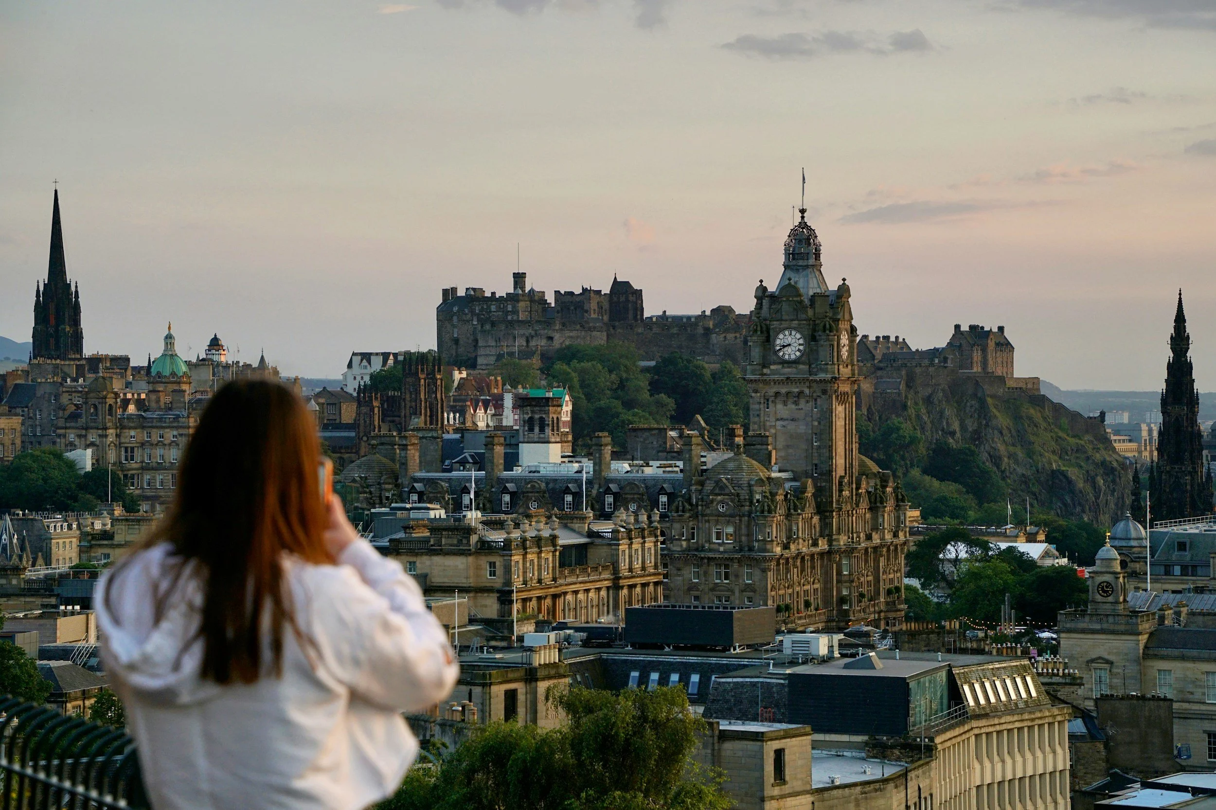 A woman with brown hair wearing a white jacket is looking at the Edinburgh cityscape with historic buildings, clock towers, and a castle on a hill during sunset.