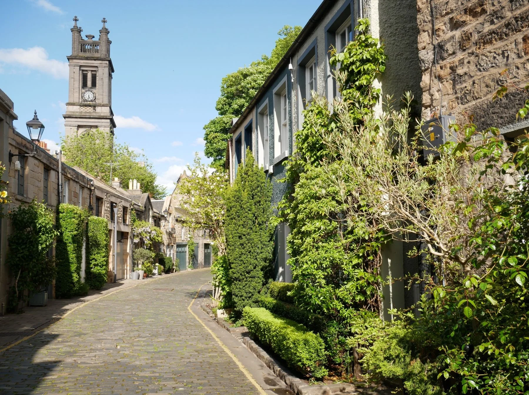 A cobblestone street in Edinburgh lined with green bushes and trees, with residential buildings on one side and a clock tower in the background under a blue sky with some clouds.
